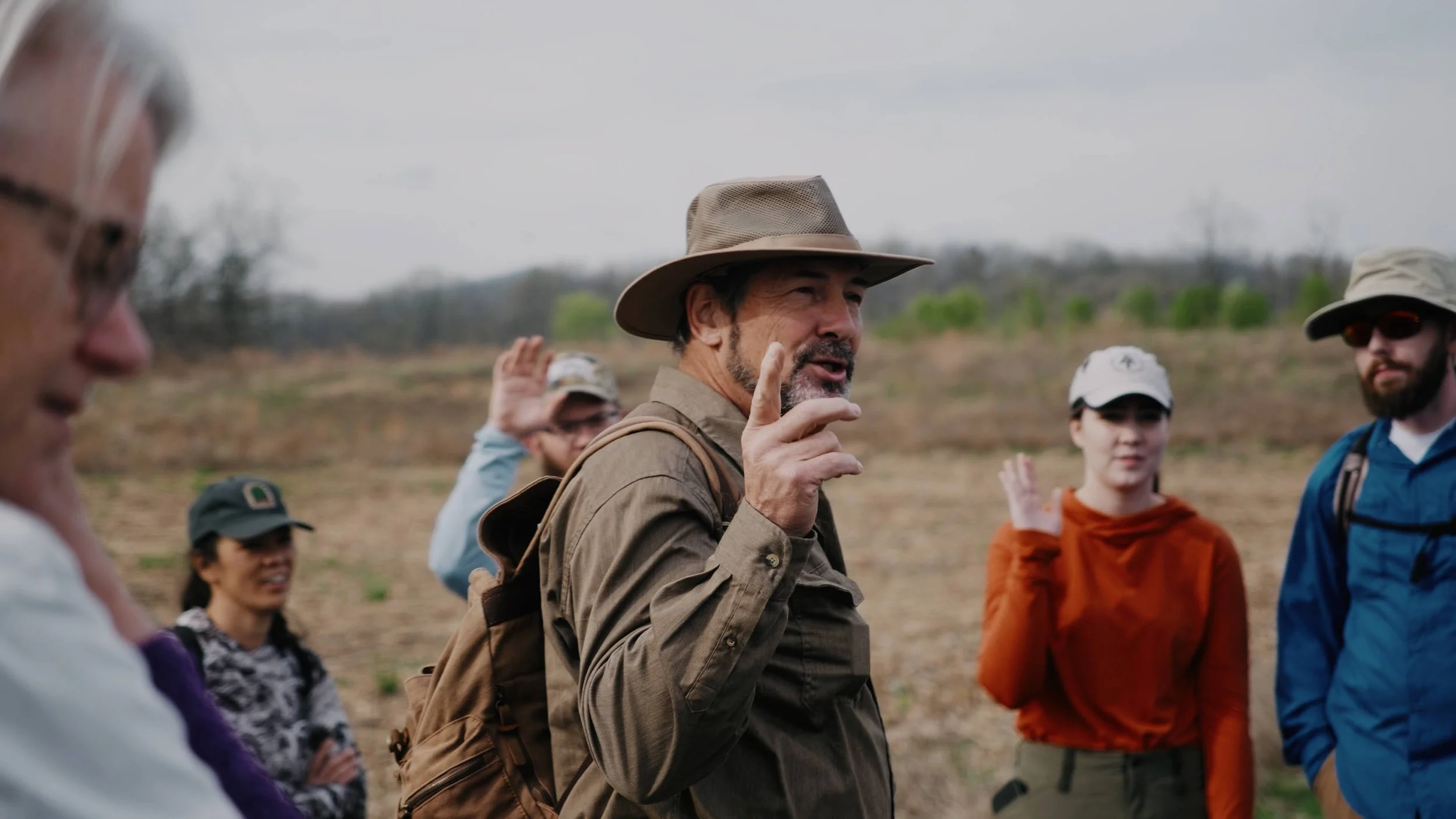 A man in a tan hat and jacket leading a group on a nature hike outdoors, with trees and a cloudy sky in the background.