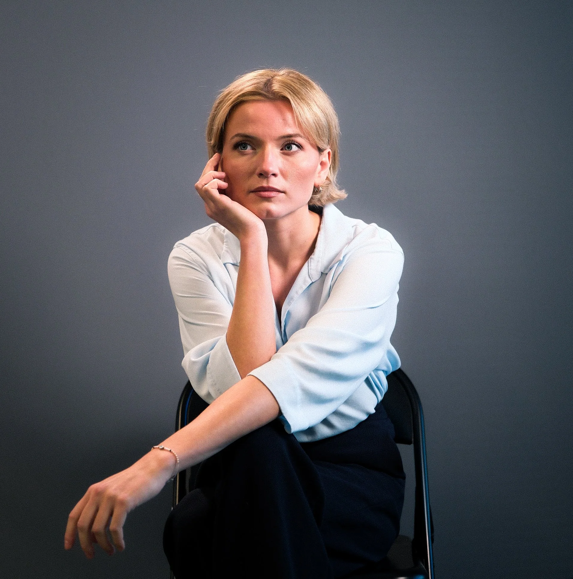 A woman with short blonde hair wearing a white shirt, sitting with her head resting on her hand against a gray background.
