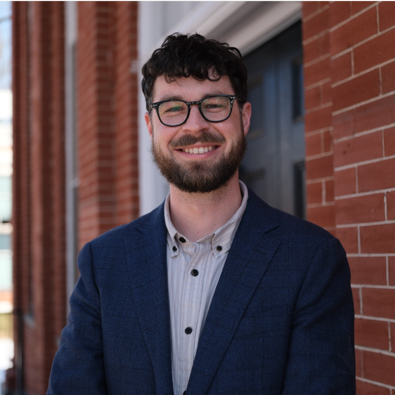 A man with dark curly hair, glasses, and a beard, smiling outdoors in front of a brick building, wearing a blue blazer and a light-colored shirt.