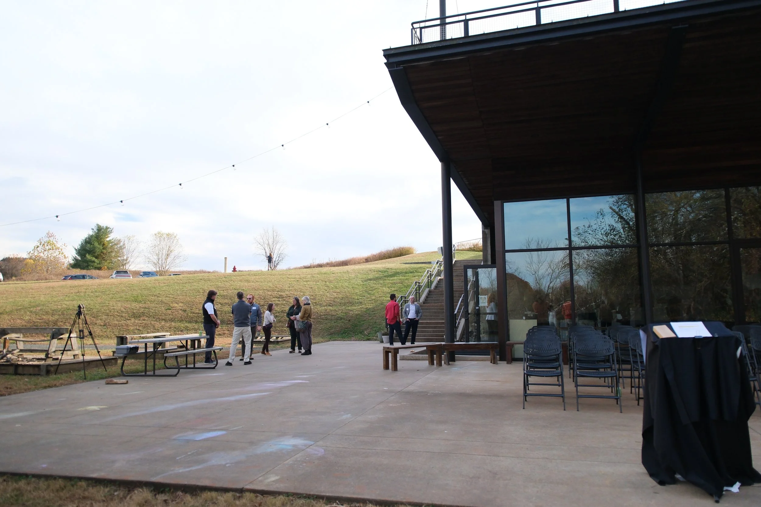 Group of people standing outside a modern building with large windows, with a grassy hill and trees in the background, and string lights hanging above.