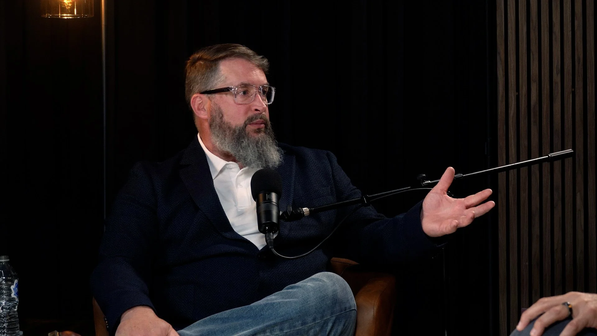 A man with glasses and a beard is sitting and gesturing with his right hand while speaking into a microphone. He is wearing a dark blazer and a white shirt, and he appears to be in a discussion or interview setting.