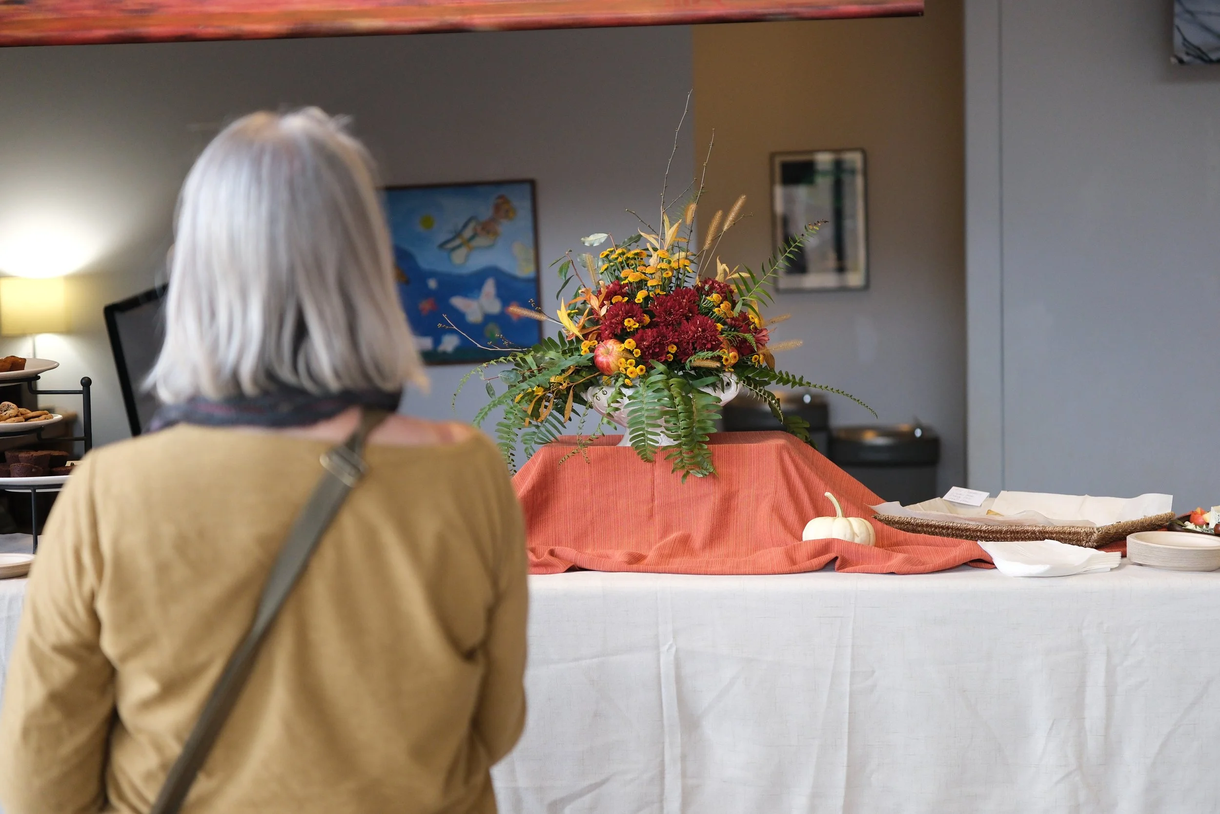 A woman with gray hair and a beige jacket looking at a floral arrangement on a table covered with a red cloth inside a room.