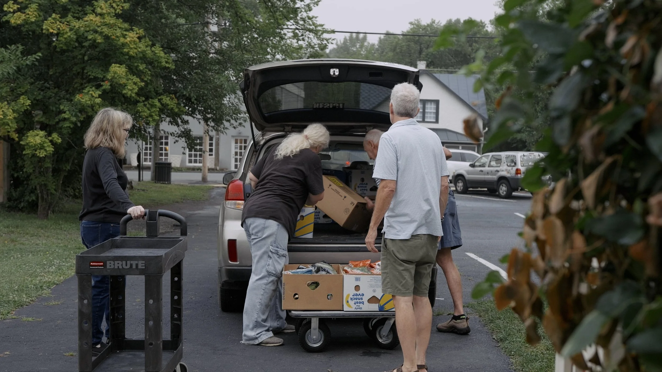 A group of five people unloading boxes from the back of a silver SUV in a parking lot surrounded by trees and parked cars.