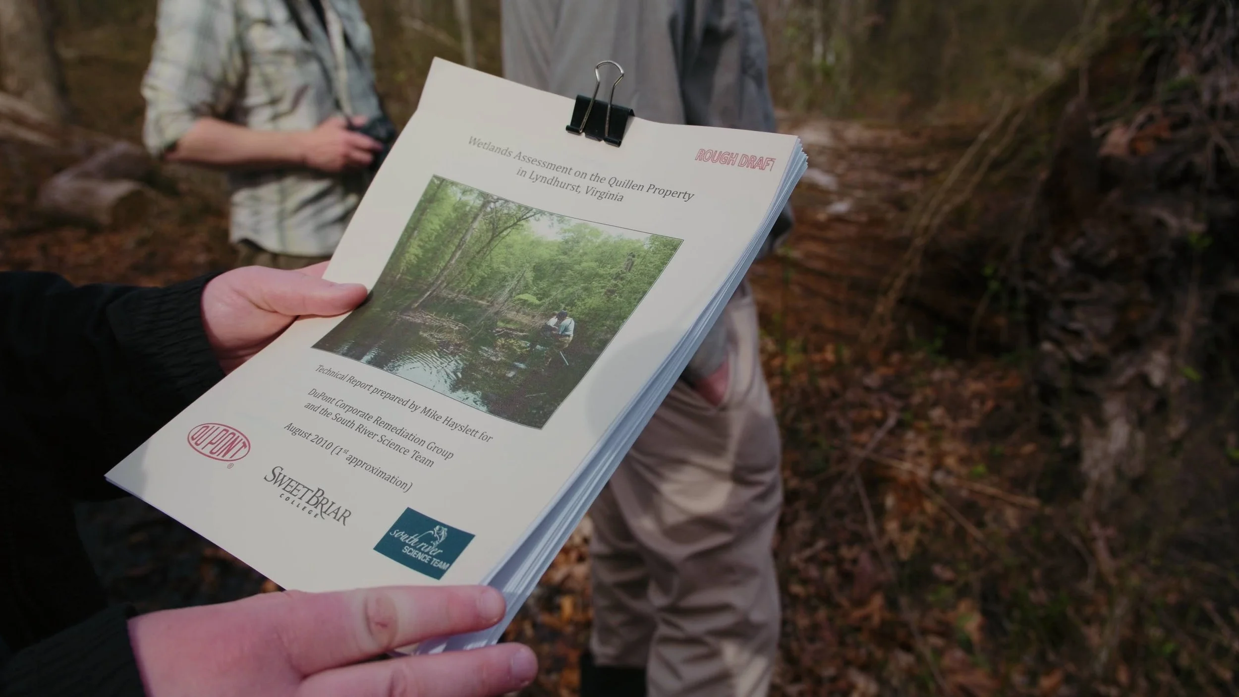 Person holding a document titled "Wetlands Assessment on the Quillen Property in Lyndhurst, Virginia" with a photo of a person fishing in a creek surrounded by trees, and another person standing nearby in a wooded area.