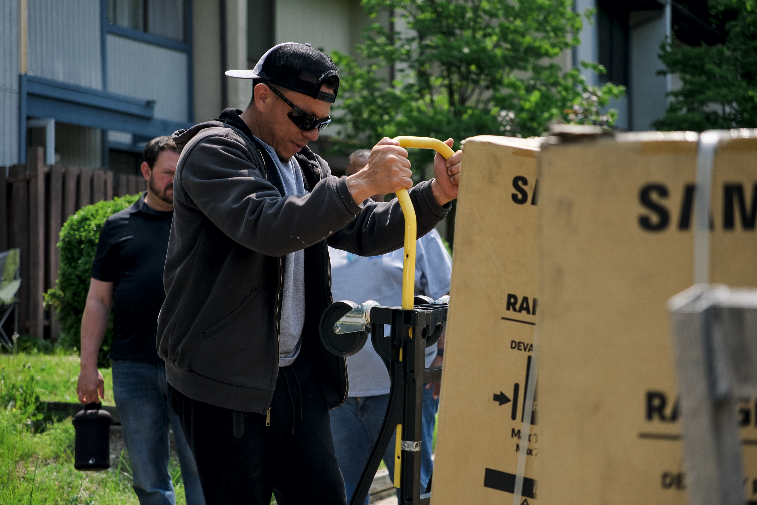A man wearing a black baseball cap, sunglasses, and a black jacket is handling a yellow tool next to a large cardboard box with black text, outdoors on a sunny day with green trees and a building in the background.