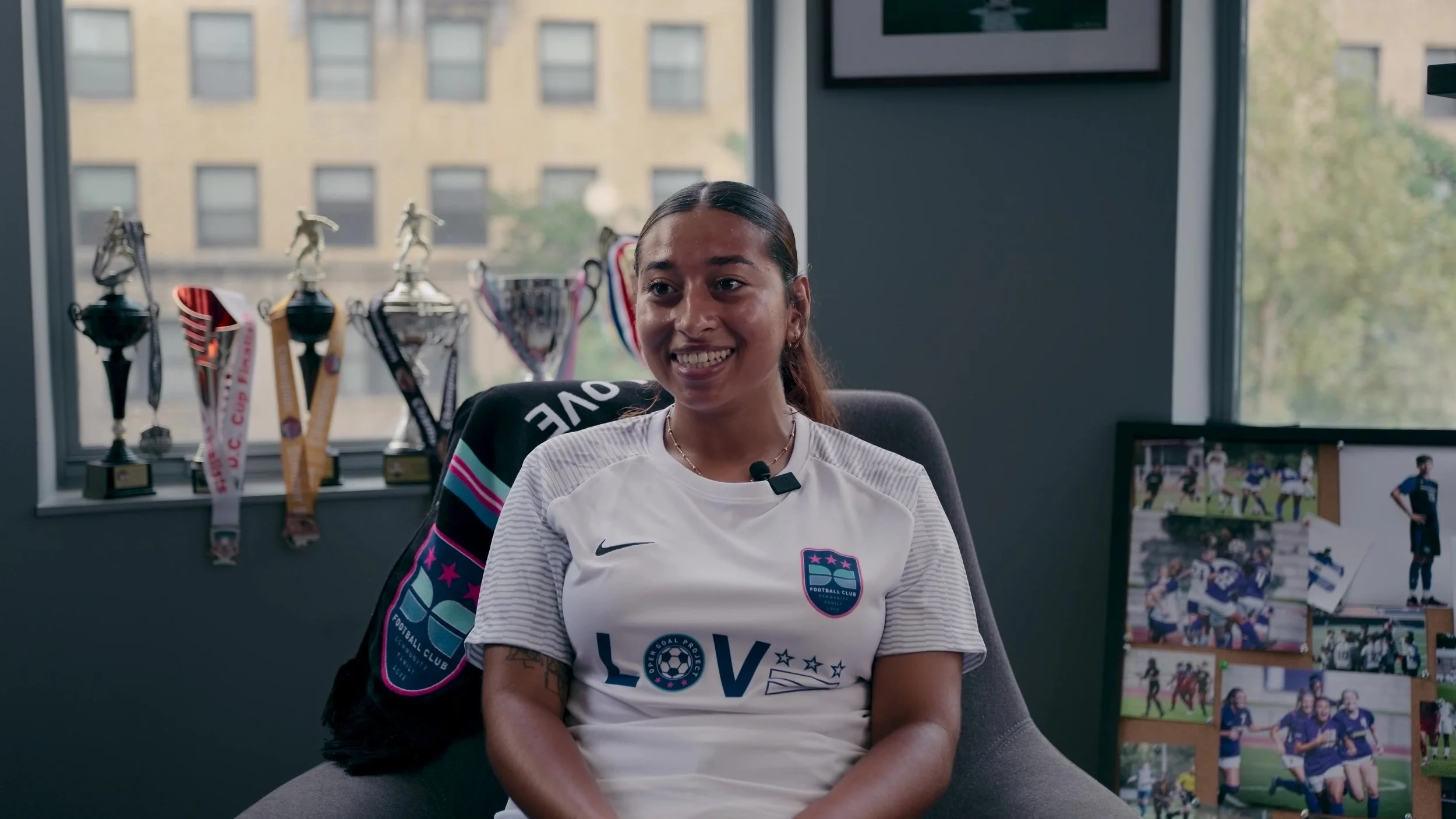 A young woman sitting in an office chair, smiling, wearing a white soccer jersey with a logo, with a window behind her displaying trophies and medals on a shelf.