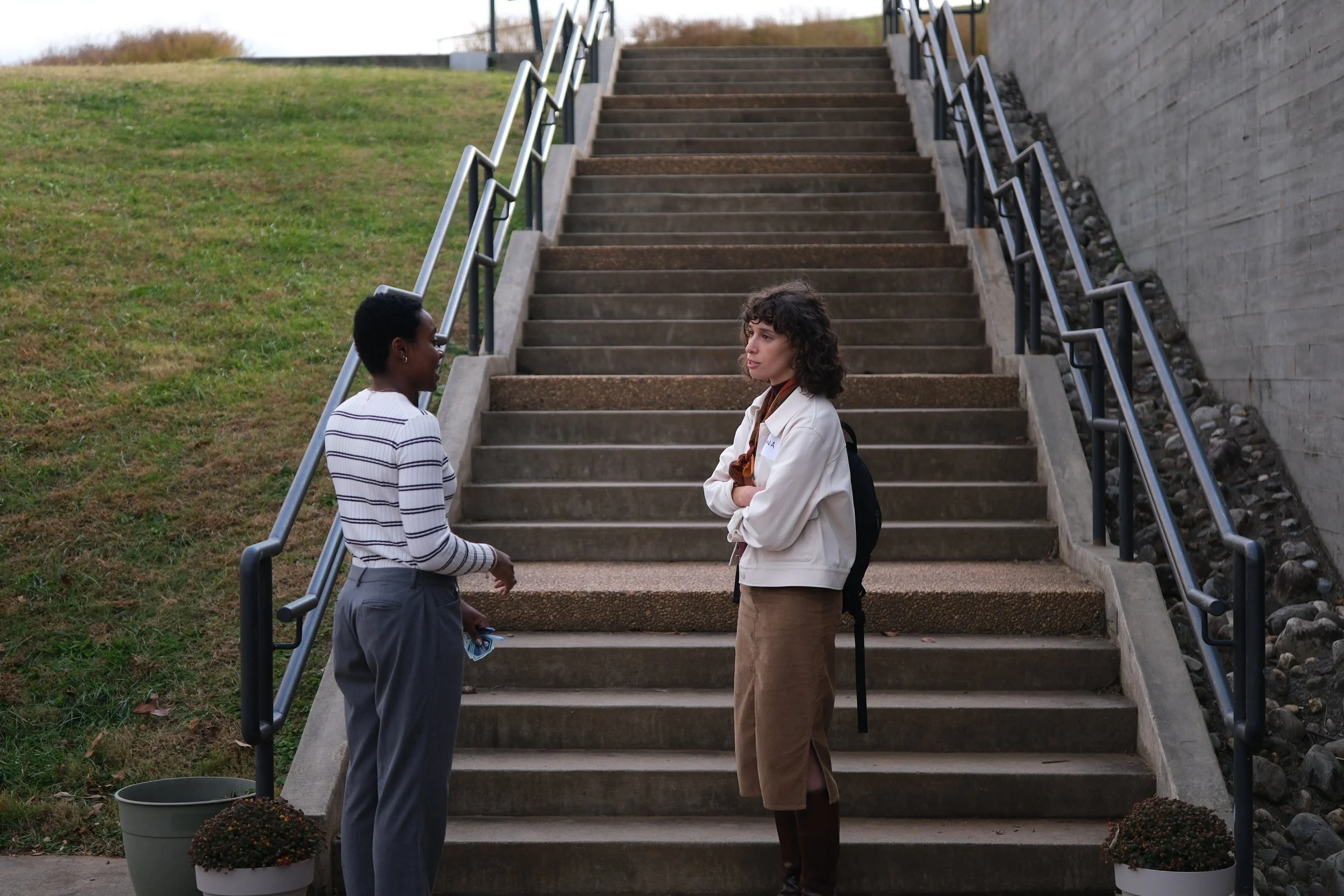 Two women stand on an outdoor concrete staircase engaged in conversation. One woman is wearing a white shirt with black stripes and gray pants, while the other is in a white jacket and brown pants with her arms crossed. The staircase has black handra