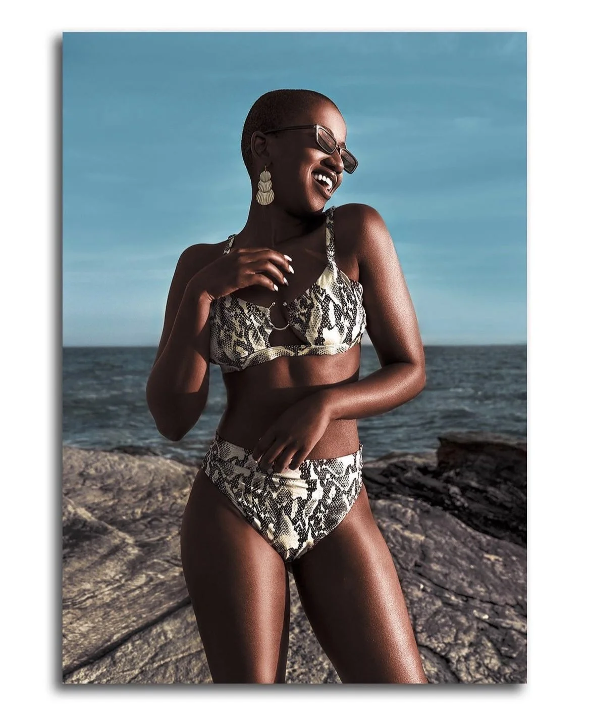 A woman in a snakeskin bikini standing on rocks near the ocean, smiling and laughing, wearing sunglasses and earrings under a clear blue sky.