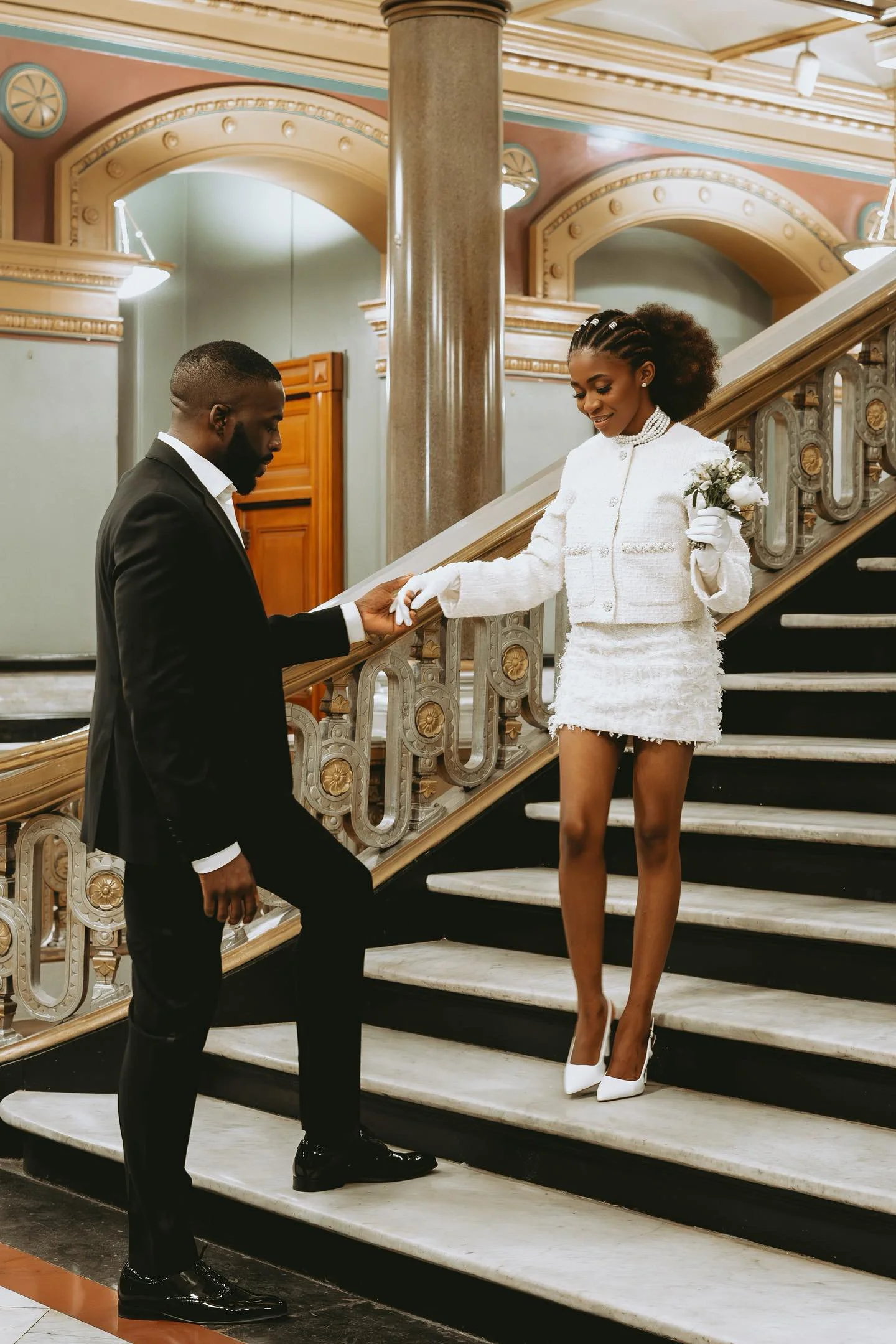 A man proposing to a woman on a staircase in a grand, ornate building. The woman is dressed in white, holding a small bouquet, and the man is in a black suit. The woman is smiling as she receives the ring.