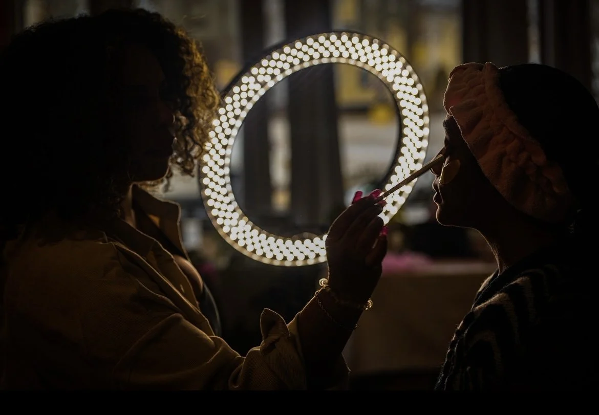 Silhouettes of two women, one applying makeup to the other in front of a lit mirror at dusk or nighttime.