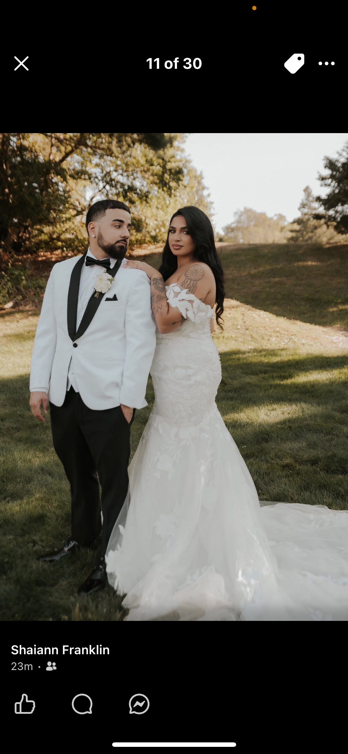 A bride and groom in wedding attire outdoor in a park. The groom is wearing a white tuxedo with a black bow tie, and the bride is in a white wedding gown with lace details and tattoos on her arms.