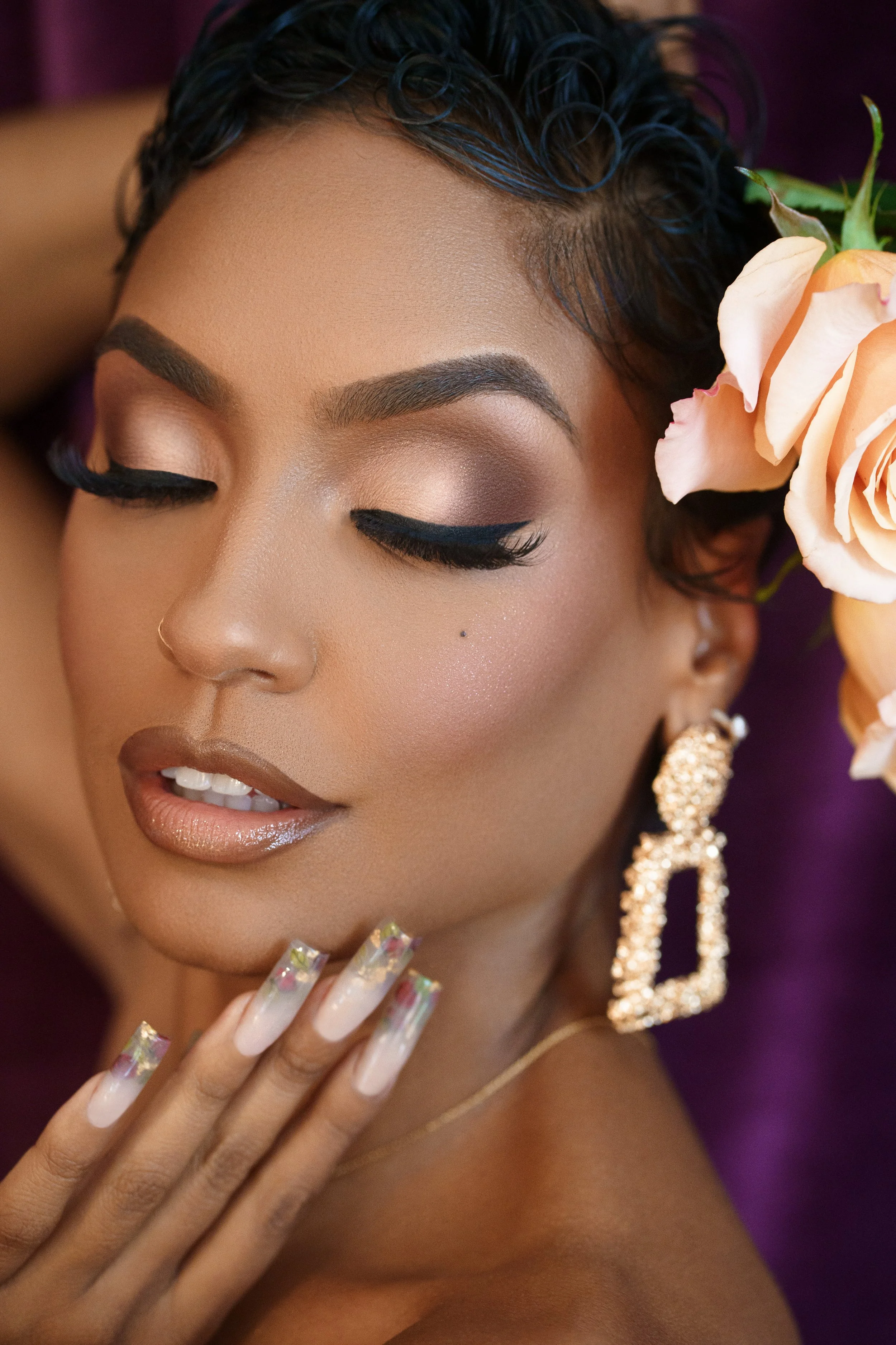 Close-up of a woman with flawless makeup, dark curly hair, a peach-colored rose near her face, large gold earrings, and manicured nails, with her eyes closed.