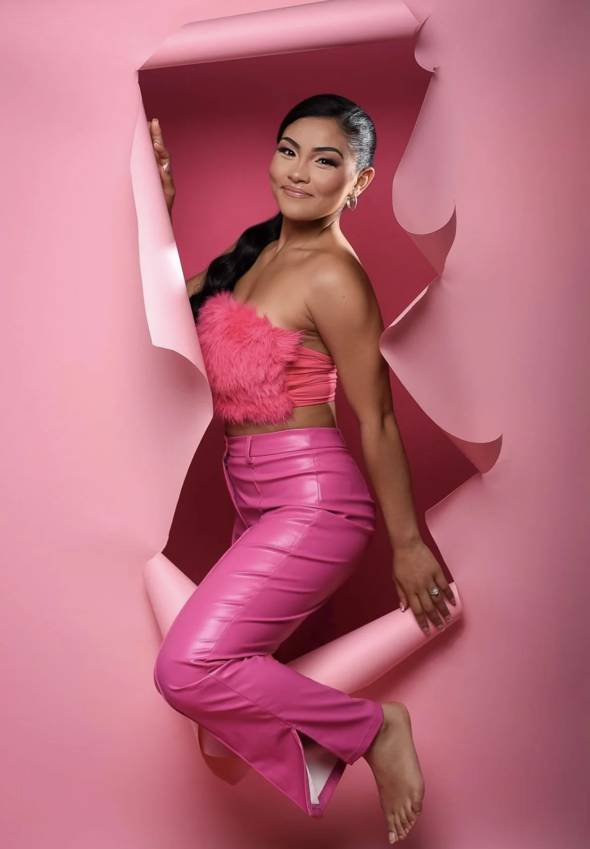A woman with black hair in a side-swept ponytail, winking, standing inside a pink paper cutout with ripped edges, wearing a pink furry top and high-waisted pink pants, with a pink background.
