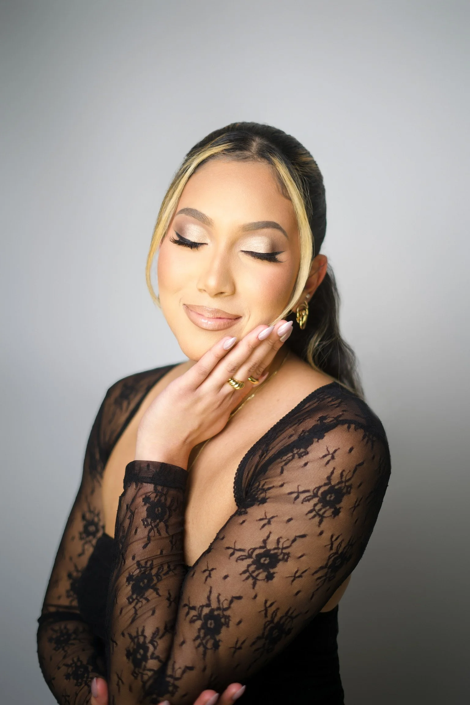 A woman with blonde highlights in her dark hair, wearing makeup with a natural lip and eye shadow, dressed in a black lace top, with her eyes closed and a serene expression, touching her chin with her right hand.