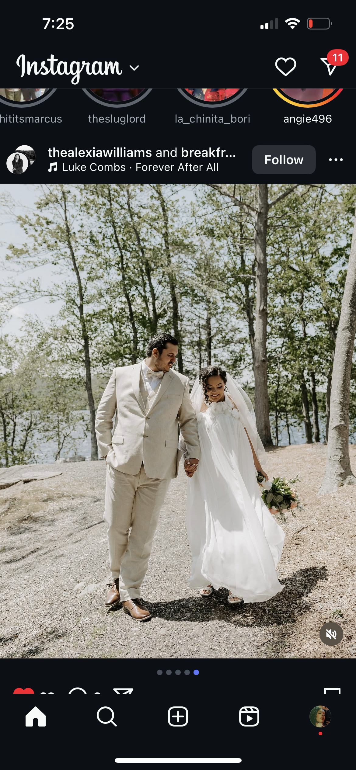 A bride and groom walking hand in hand outdoors, smiling, during a wedding photoshoot in a wooded area with trees and a body of water in the background. The bride wears a white gown and carries a bouquet, the groom wears a beige suit.