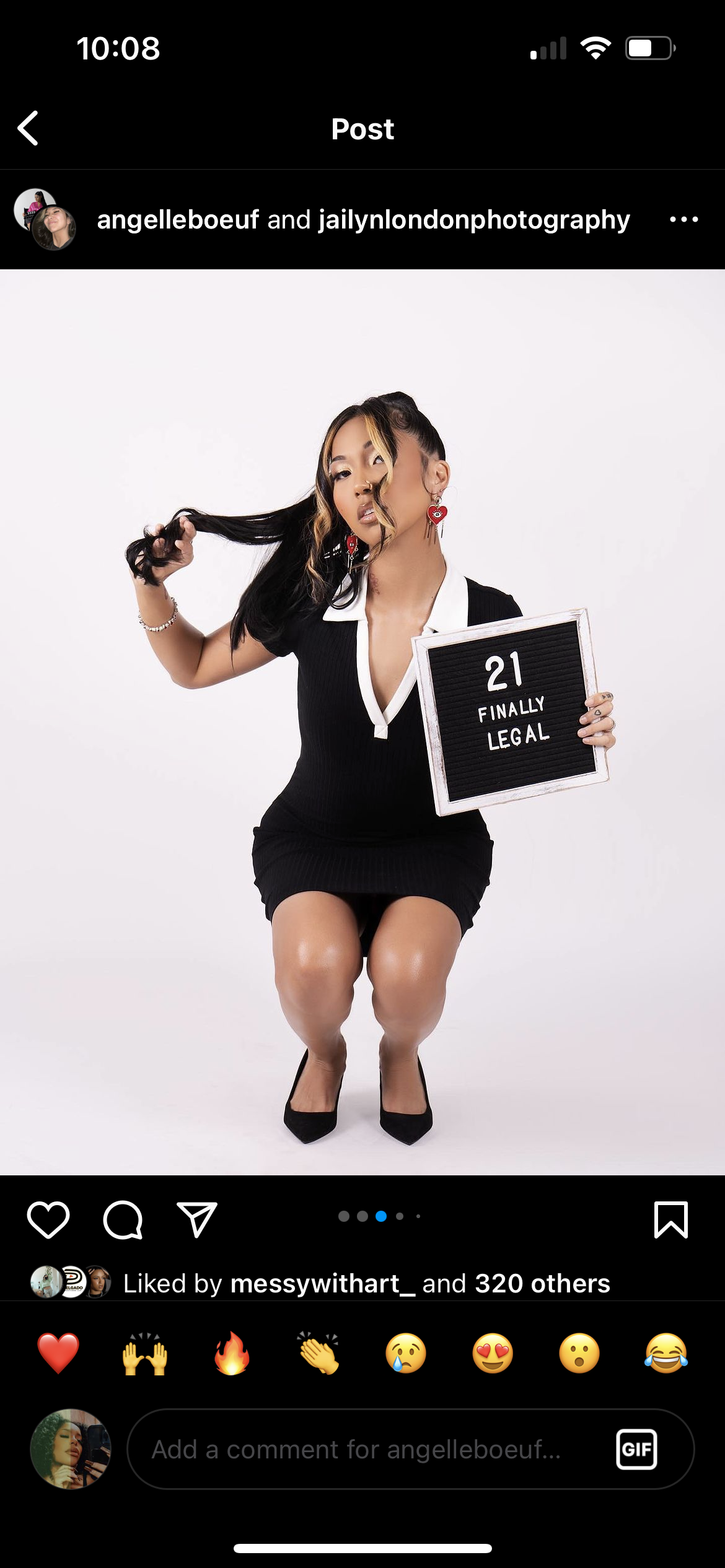 A woman with black hair and blonde streaks, wearing a black dress with white collar and heels, is holding a sign that reads '21 Finally Legal' while squatting against a plain white background.