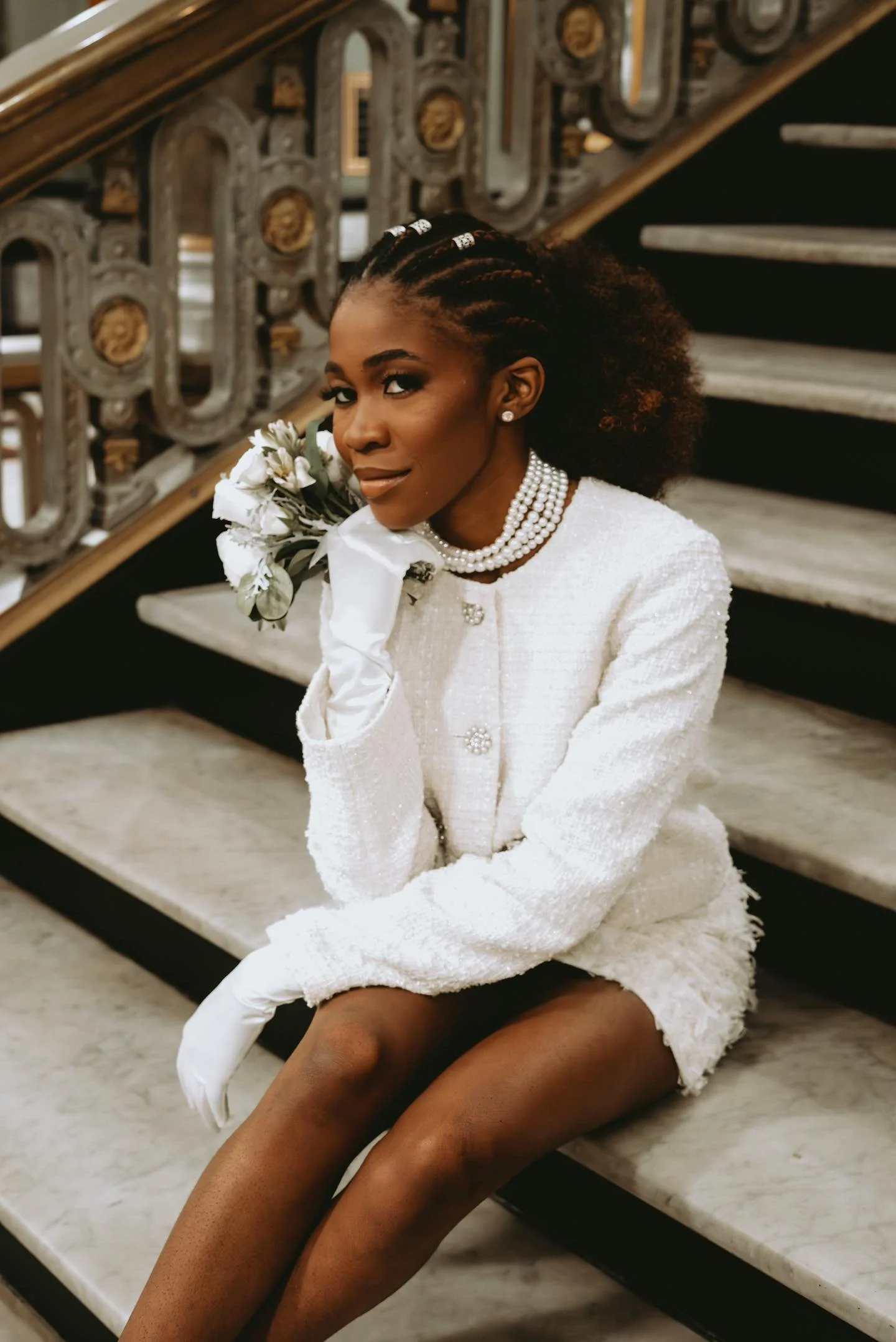 A woman dressed in a white textured jacket, pearl jewelry, and white gloves is sitting on marble stairs, holding a bouquet of white flowers, with curly hair styled in cornrows.