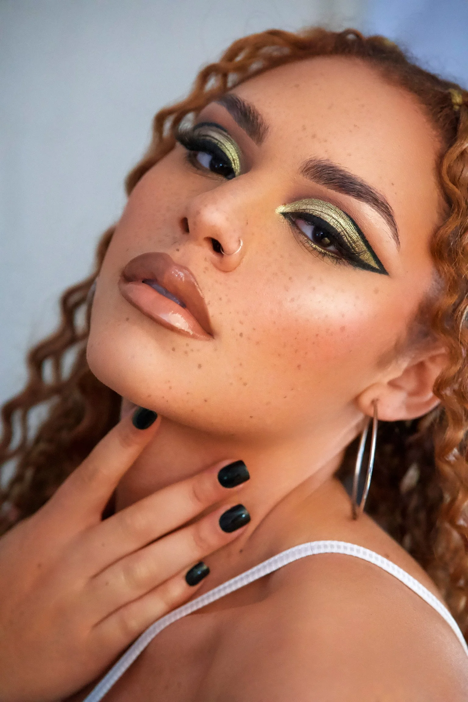 Close-up of a woman with curly hair, green eye makeup, black eyeliner, hoop earrings, and a nose ring, touching her chin with her fingers, wearing a white strap top.