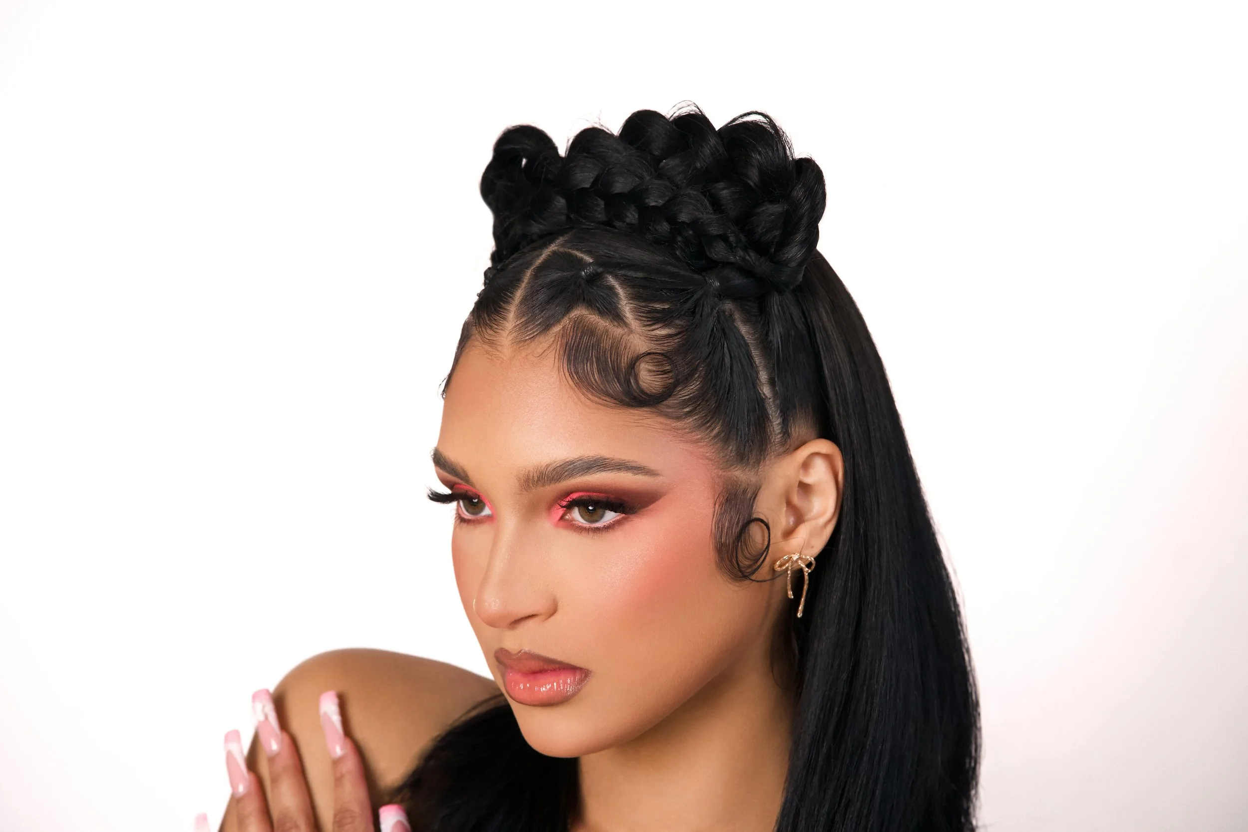 A woman with intricate braided and styled black hair, makeup, and jewelry, with a neutral background.