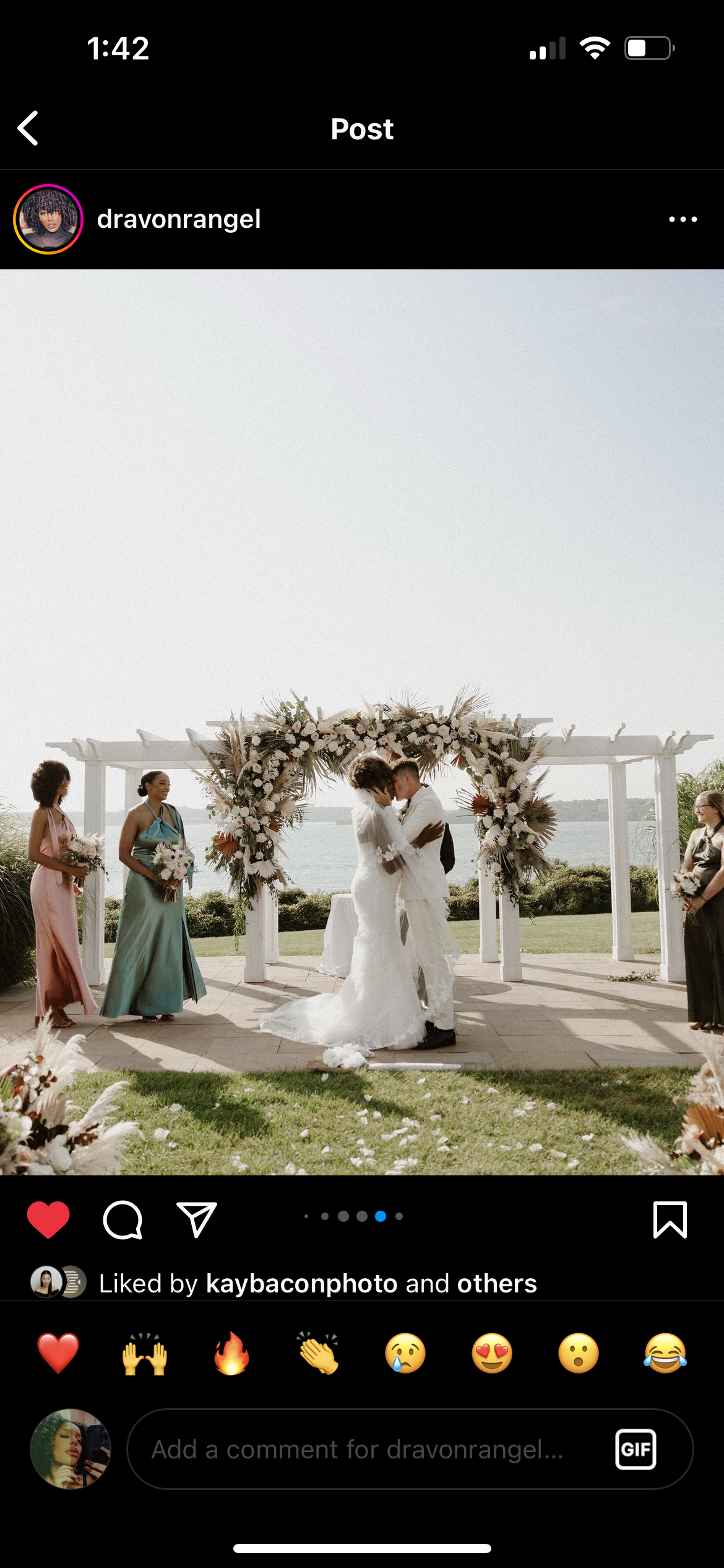 A wedding ceremony outdoors by a body of water with a floral arch, a bride and groom embracing, and women holding bouquets nearby.