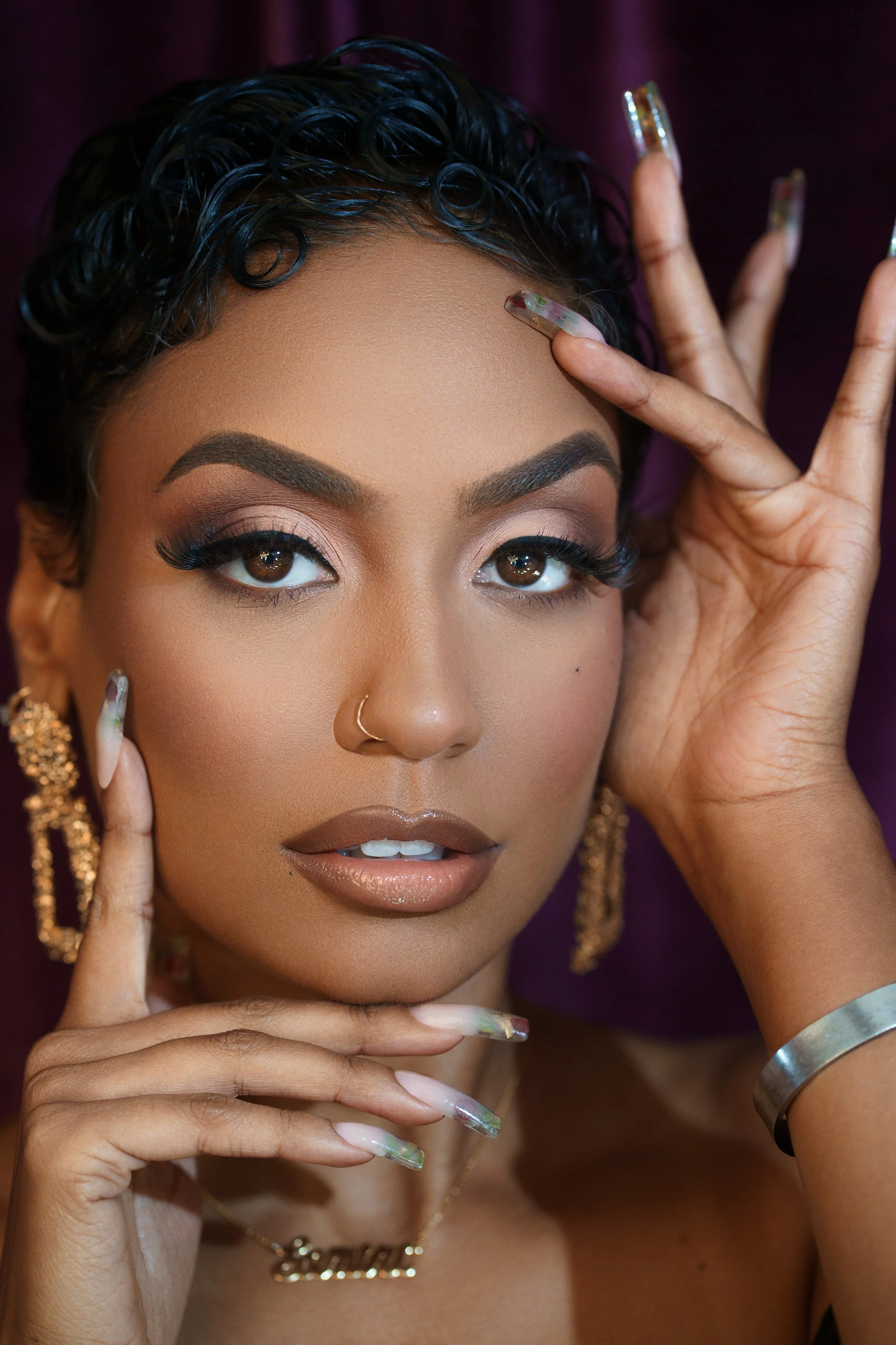 A woman with dark curly hair and makeup poses with her hands near her face, wearing gold earrings, a necklace, and a bracelet.
