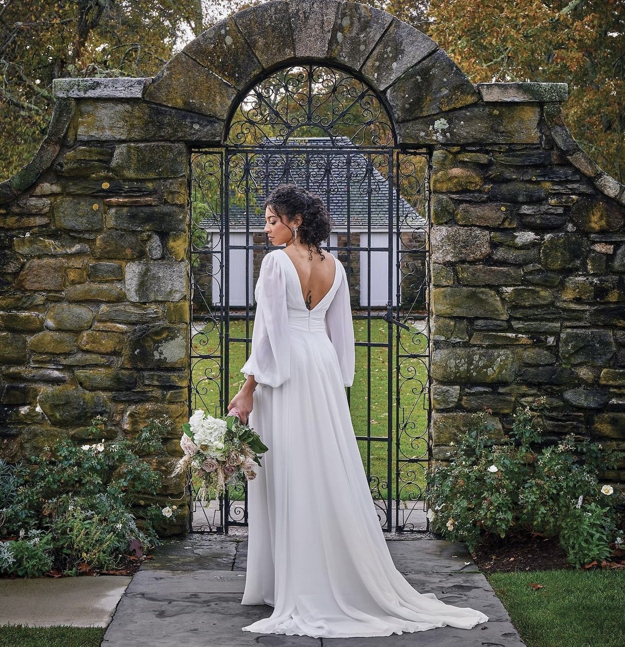 Bride in a white wedding dress holding a bouquet standing in front of an old stone and wrought iron gate.