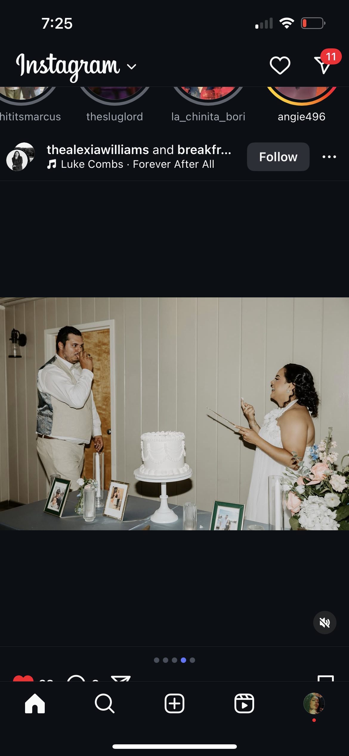 A bride and groom celebrate at a wedding reception, with the bride smiling and holding a tablet, and the groom touching his face with an expression of emotion, standing near a table with framed photos and a white wedding cake.