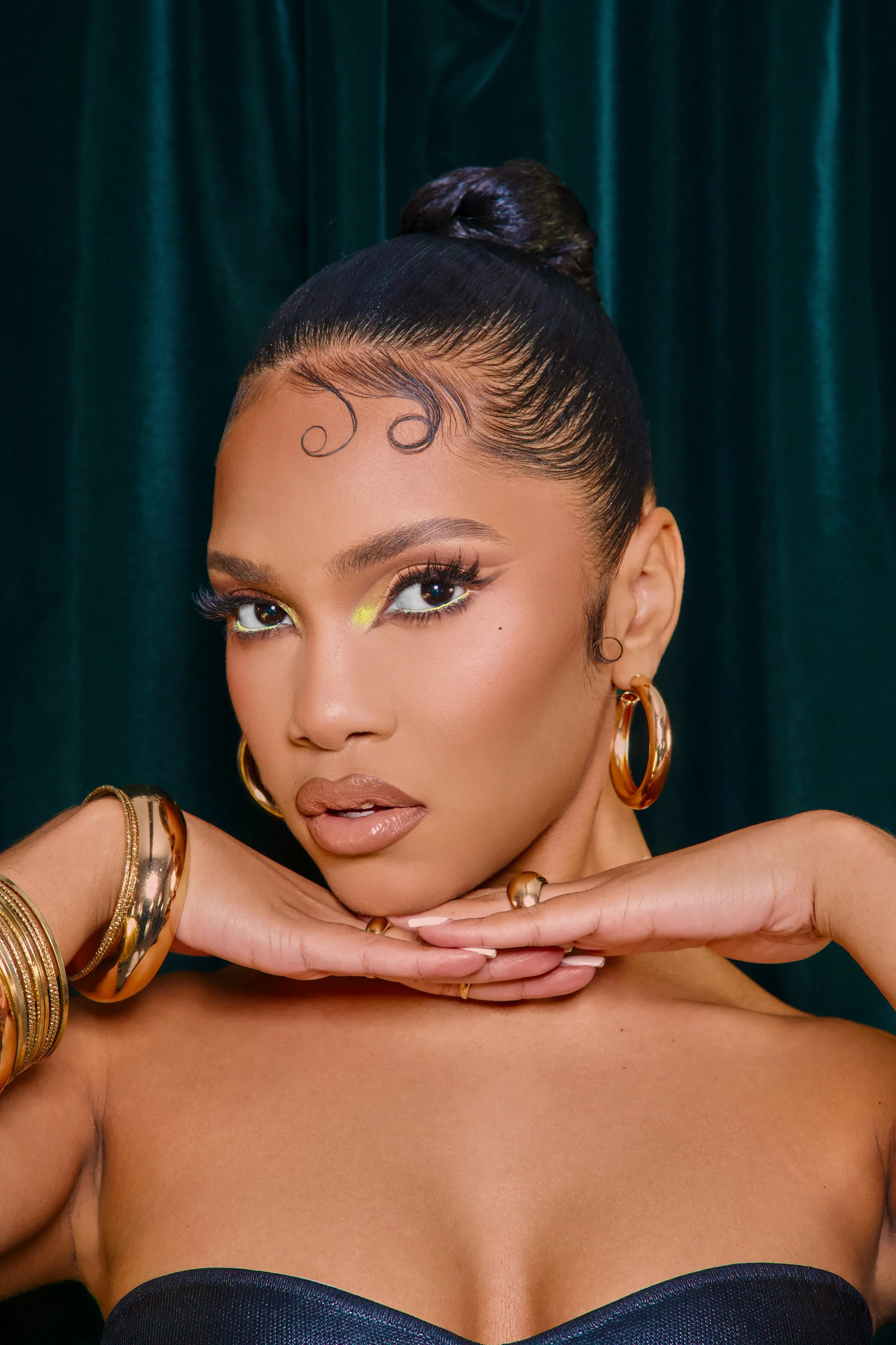 A woman with her hair in a sleek bun, wearing gold jewelry, and makeup with yellow eyeshadow, posing with hands under her chin against a dark green backdrop.