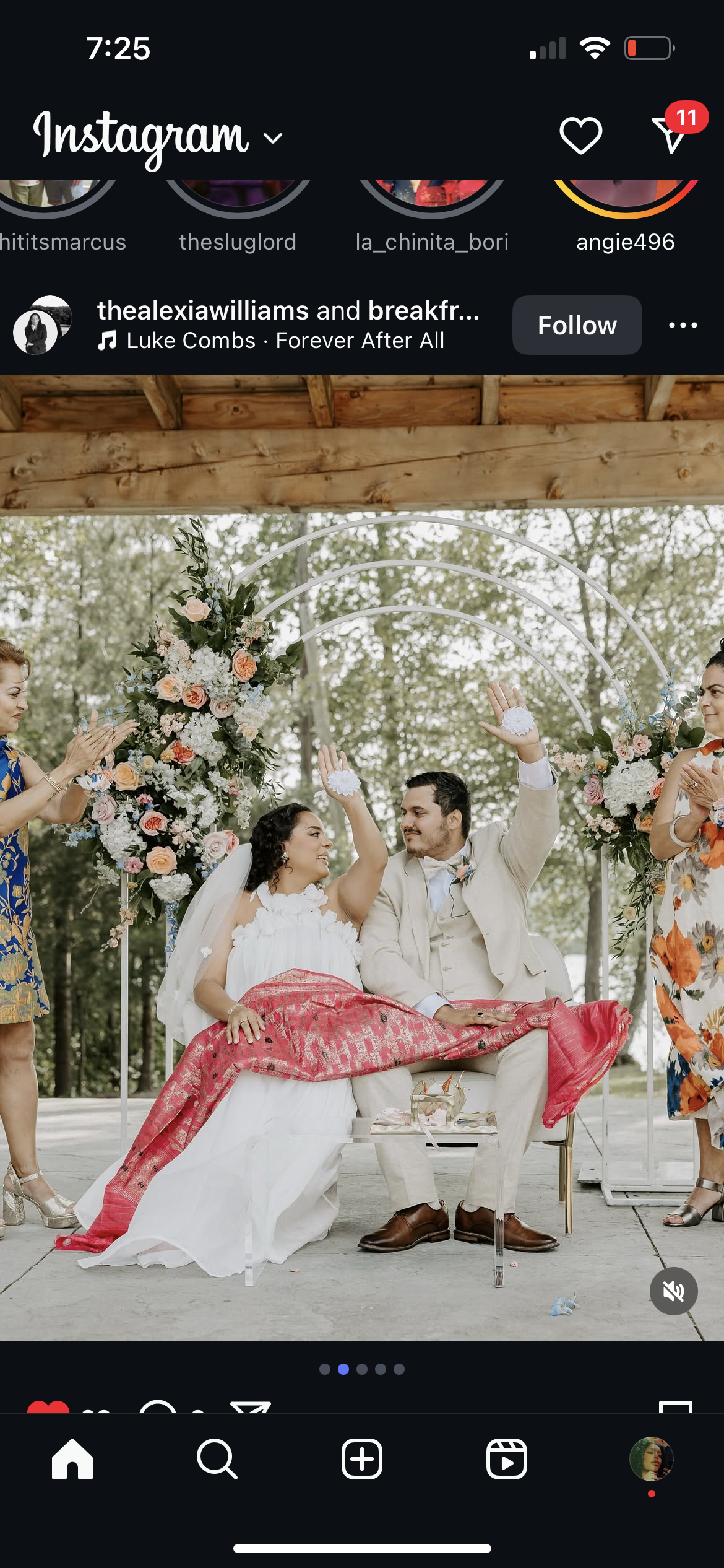 A bride and groom sitting on a bench under a decorated arch, celebrating their wedding. The bride in a white dress and the groom in a light suit share a happy moment, surrounded by flower arrangements and two women clapping on either side.