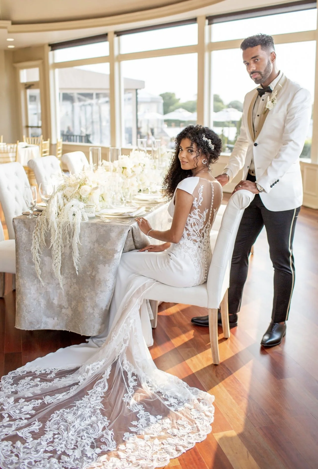 A bride and groom at a wedding reception, with the bride seated and smiling at the camera, wearing a lace wedding gown with a long train, and the groom standing behind her, wearing a white tuxedo jacket and black pants, in a bright, elegantly decorated venue with floral centerpieces and large windows.