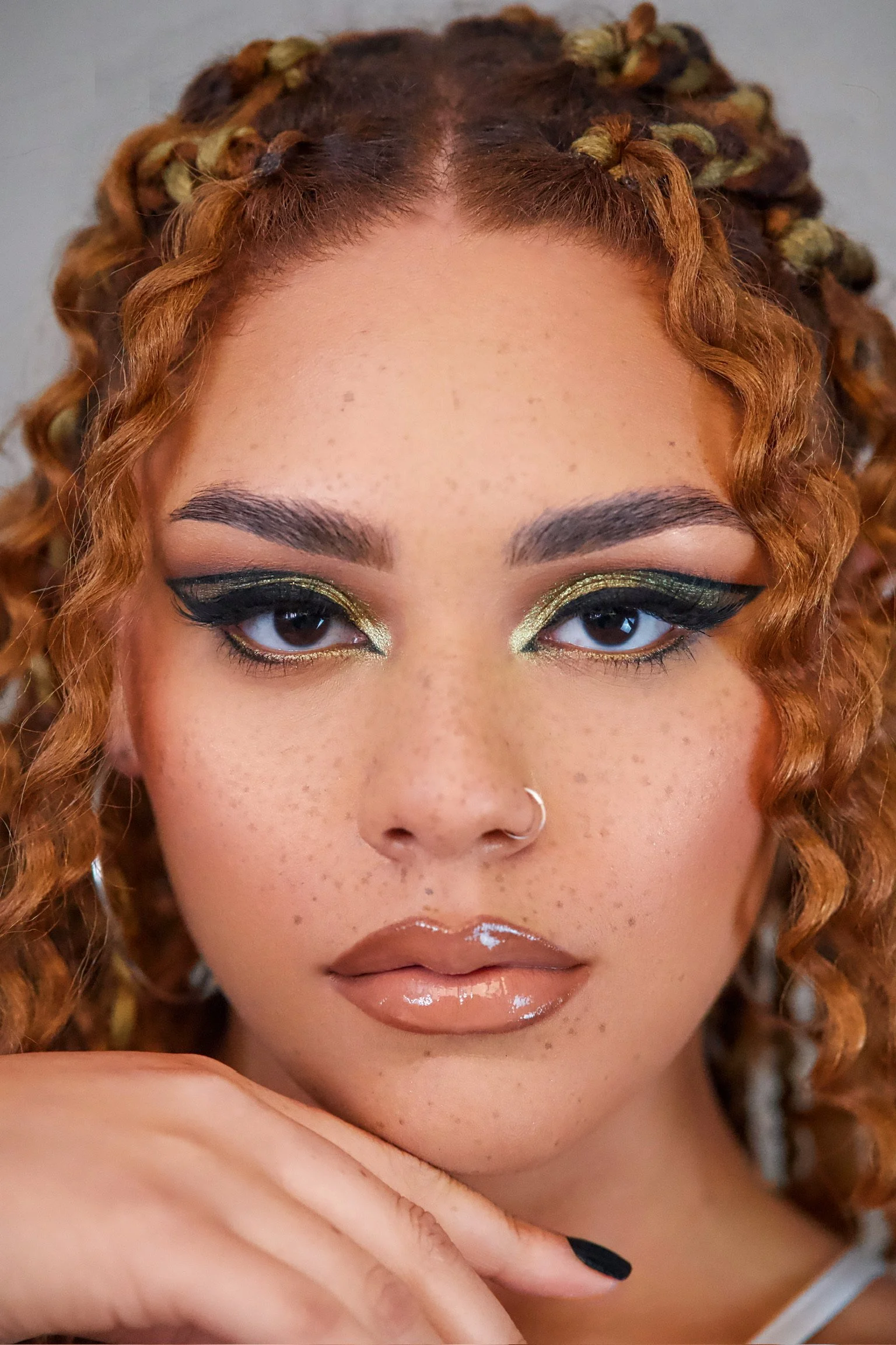 Close-up of a woman with curly hair and bold eye makeup, wearing a nose ring and glossy lips.