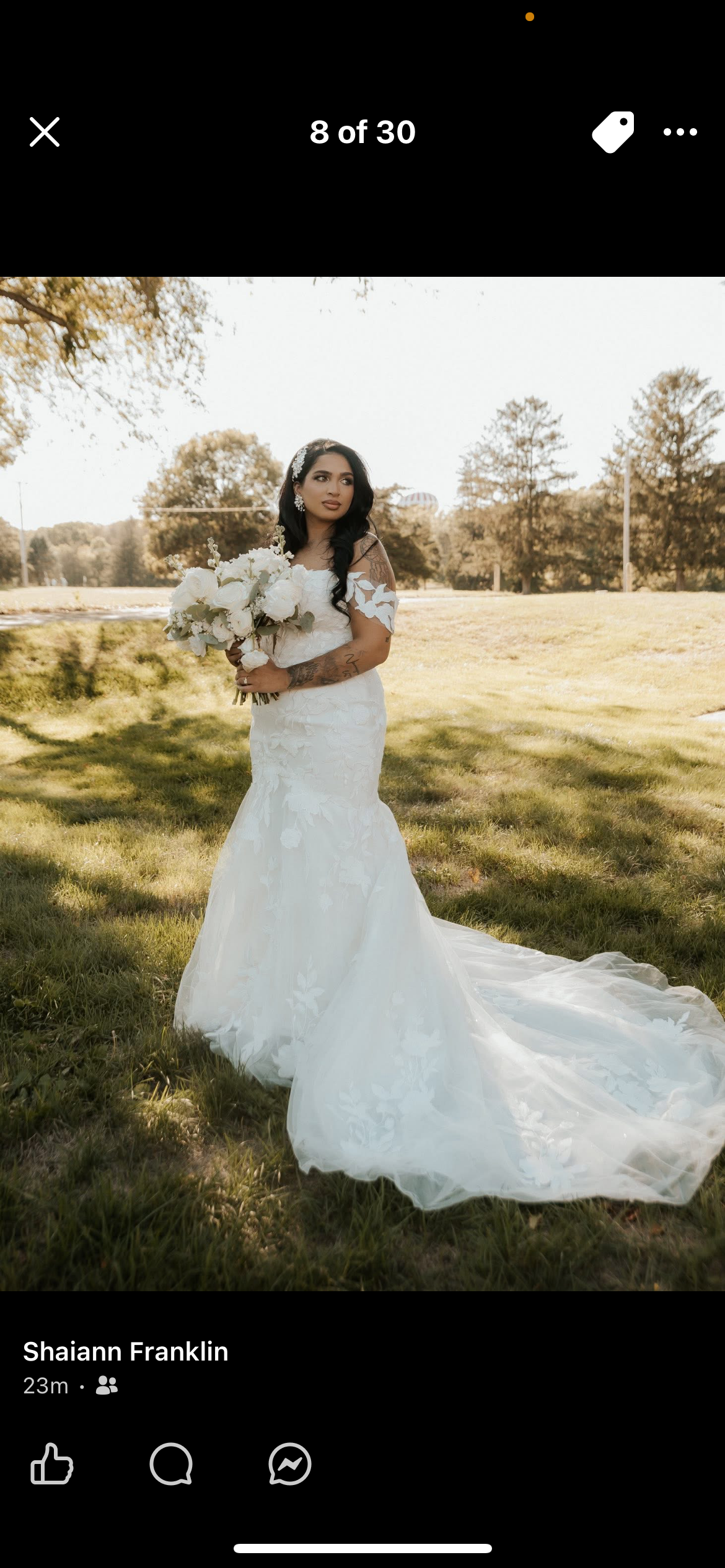 A woman in a white wedding dress holding a bouquet standing outdoors on grass with trees in the background.