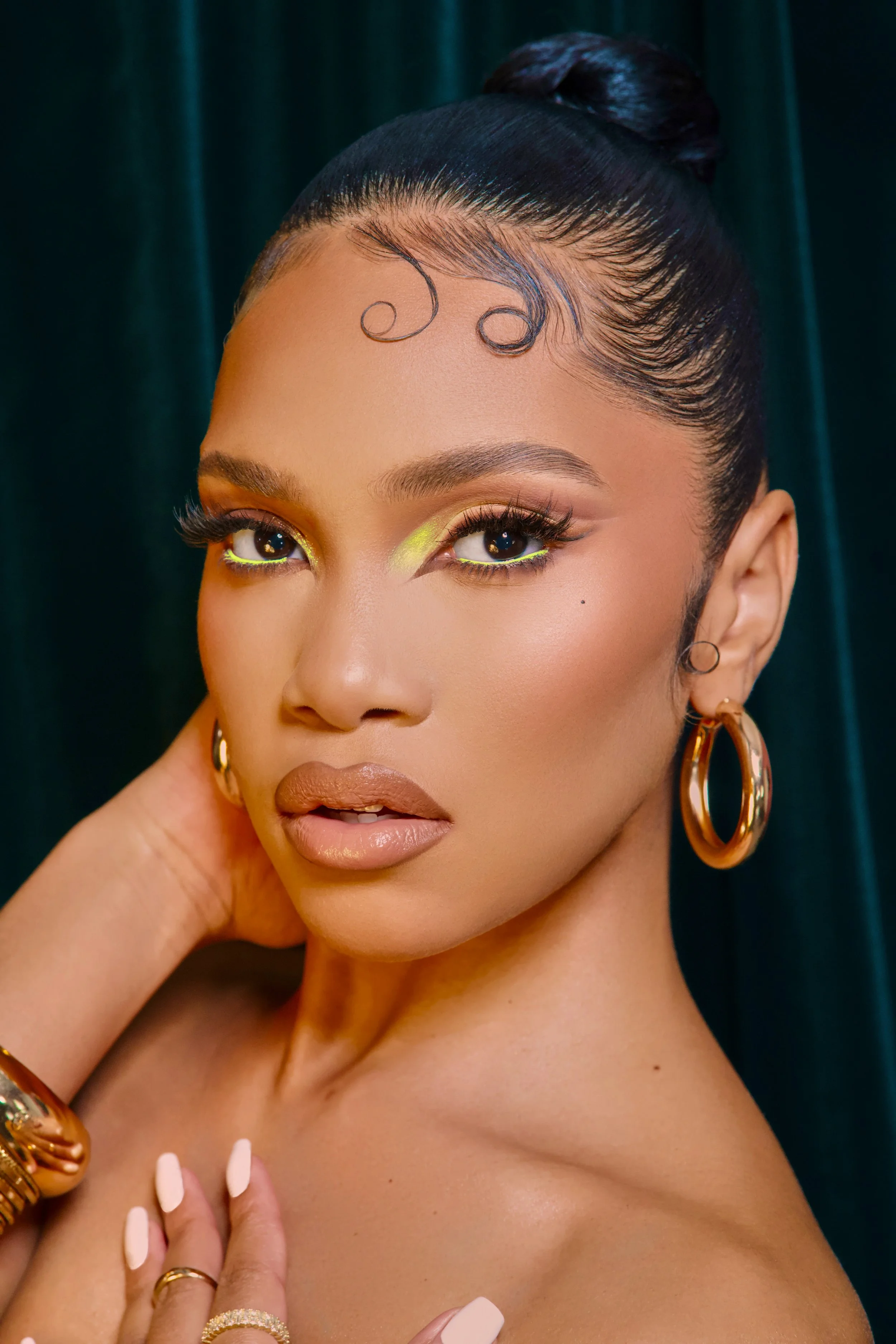 A woman with dark hair styled in a sleek bun, wearing gold hoop earrings and gold jewelry, with vibrant makeup including neon yellow eye shadow, long eyelashes, and nude lipstick, posing against a dark green background.