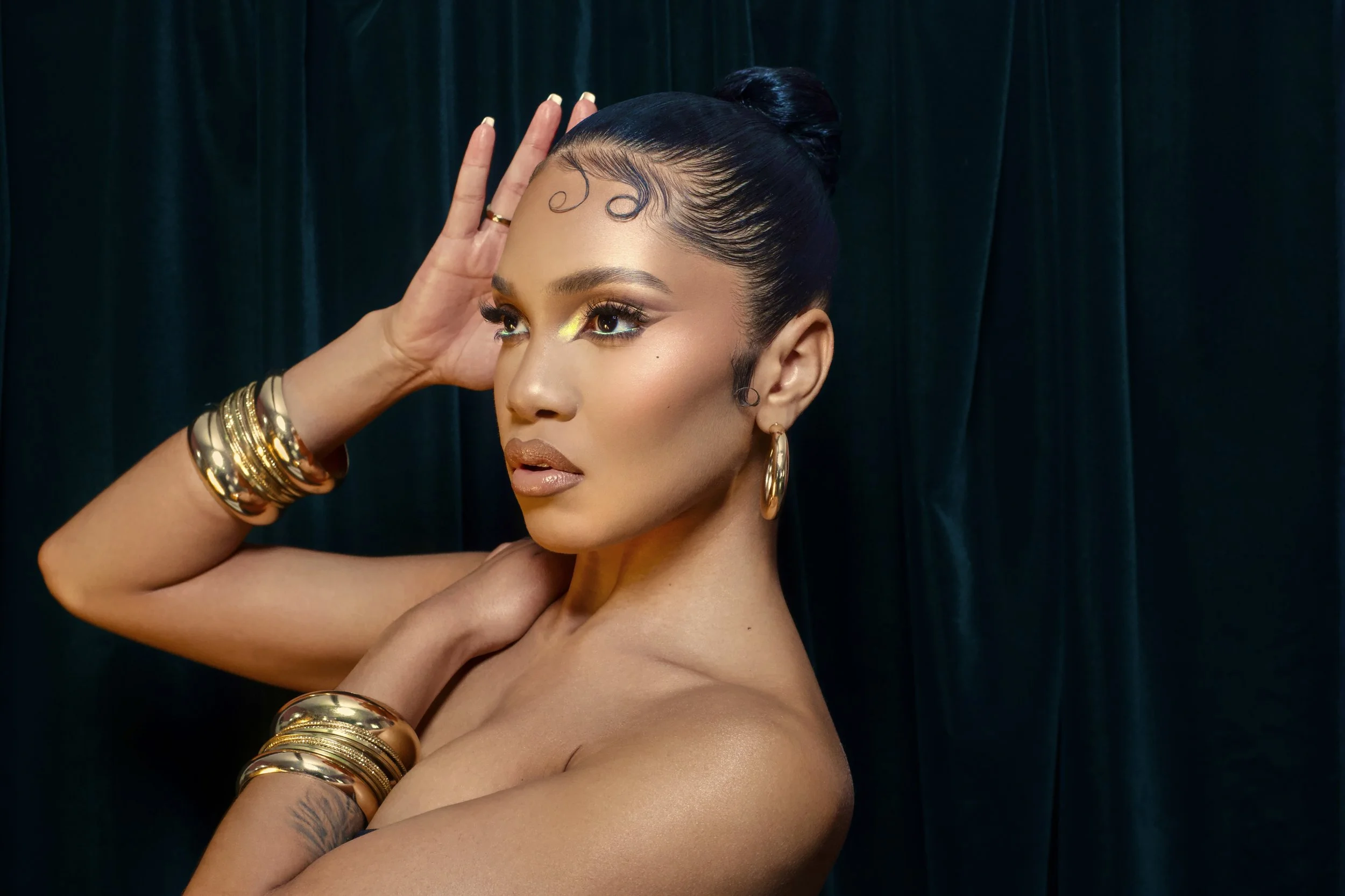 A woman with dark hair in an updo hairstyle, gold jewelry, and makeup striking a pose against a dark curtain background.