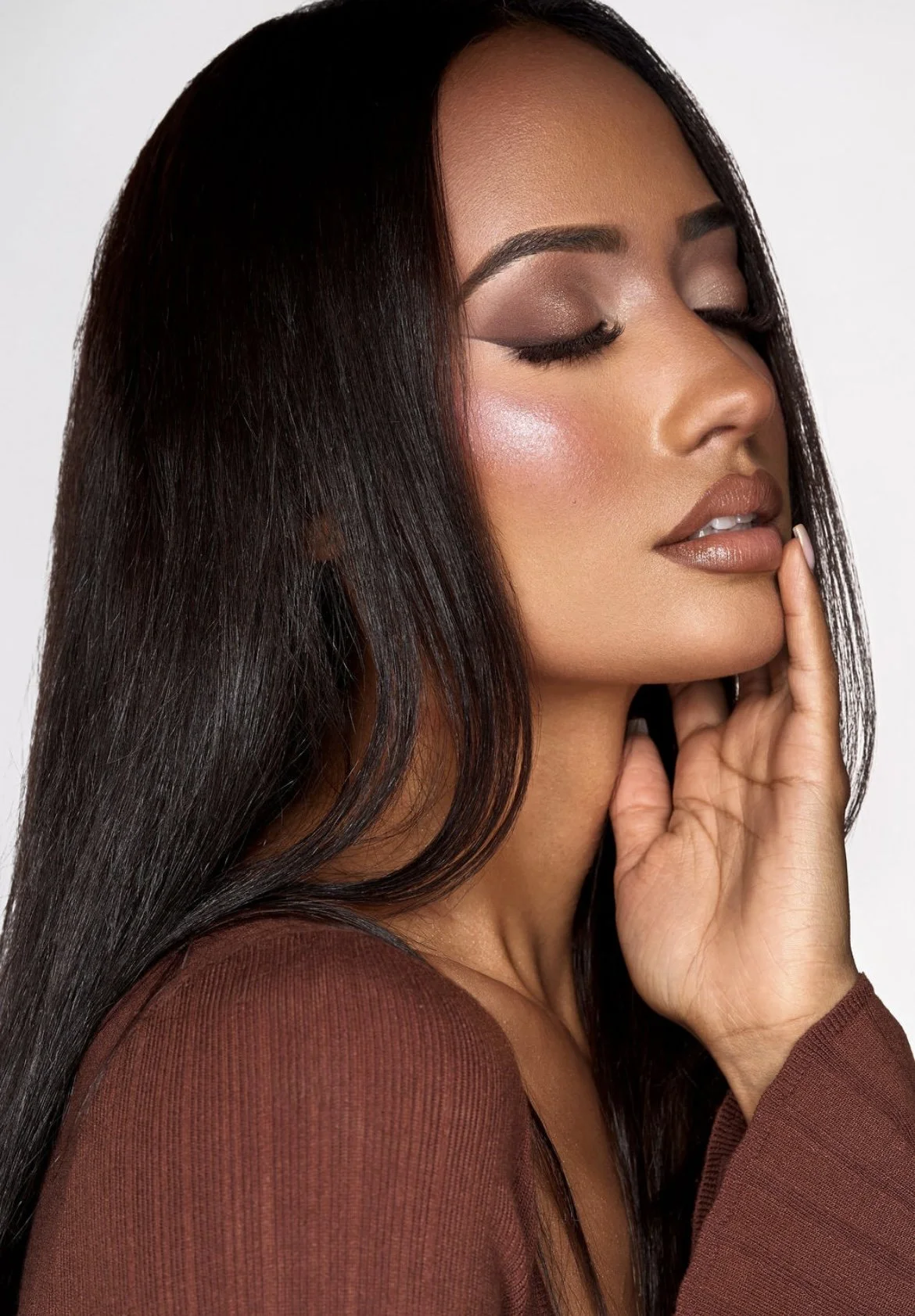 A woman with long, straight, dark hair and makeup with eyes closed and her hand touching her face.