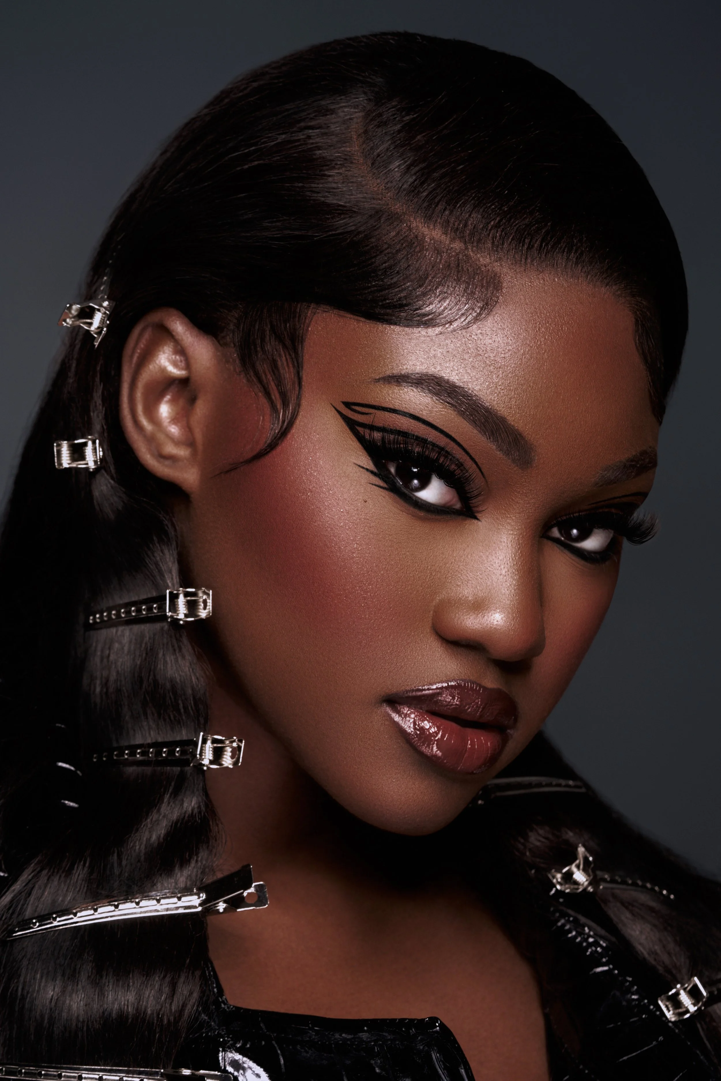 Close-up of a woman with styled hair and bold makeup, including black eyeliner, eye shadow, long lashes, and glossy lips, wearing a shiny black outfit with zipper details.