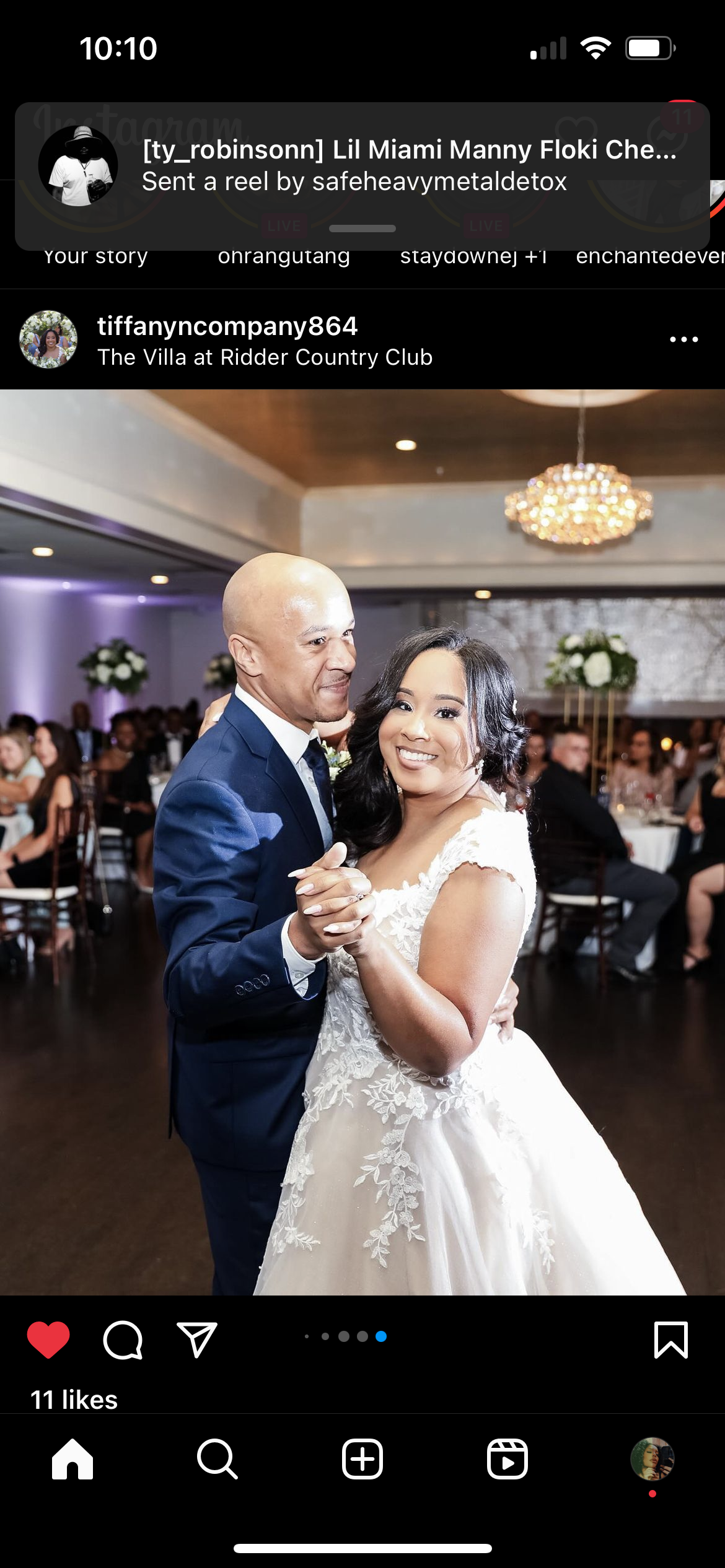 A bride and groom dancing at a wedding reception. The bride is wearing a white lace wedding gown and the groom is in a blue suit. They are smiling and holding hands. The background shows guests seated at tables and a decorated event hall with flowers