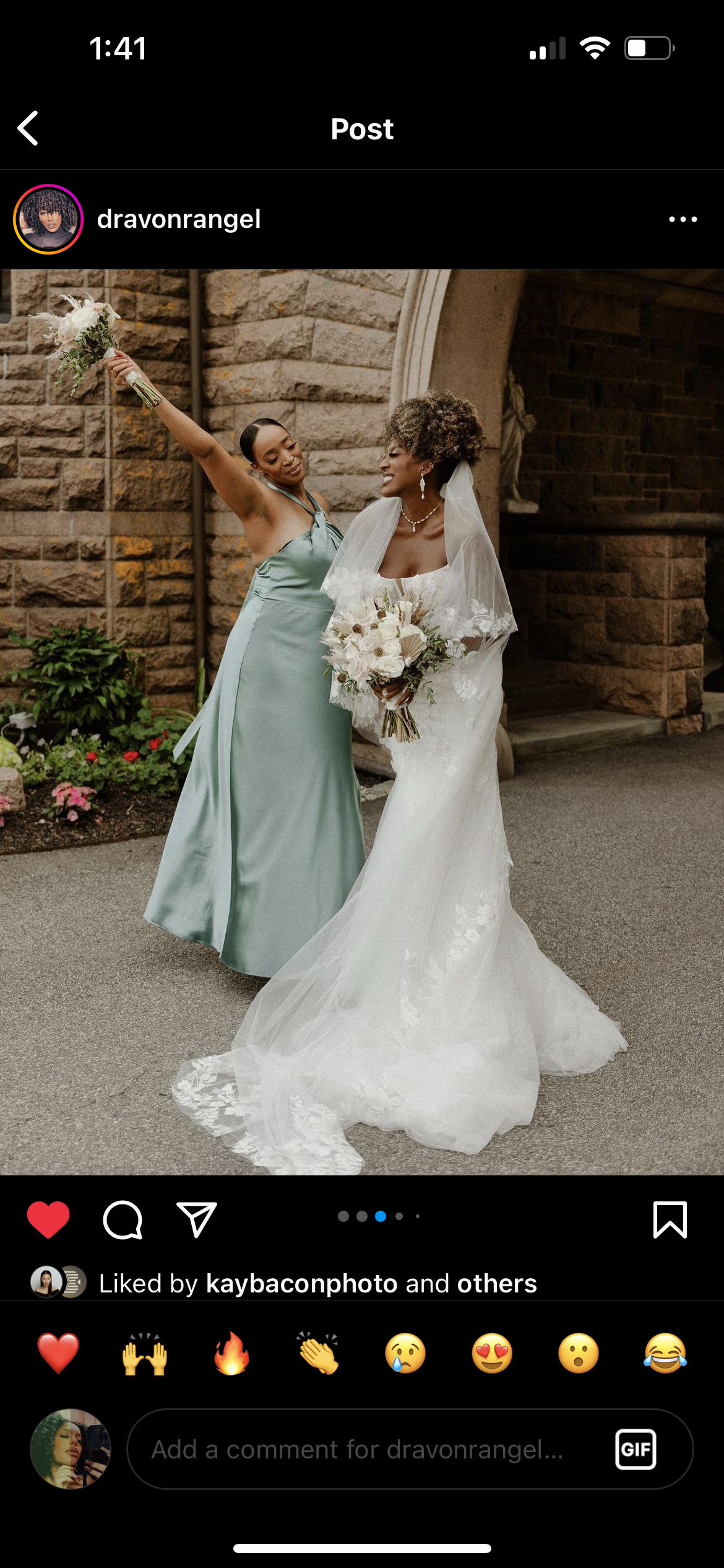A bride in a white wedding gown and veil holding a bouquet of white flowers, smiling at a bridesmaid dressed in a satin mint green gown, who is holding a bouquet in the air with one hand and touching the bride's head with the other, in front of a sto