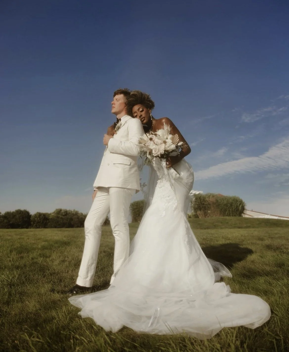 A bride and groom in wedding attire standing outdoors on a grassy field under a blue sky with sparse clouds. The bride is leaning her head on the groom's shoulder, holding a bouquet, while the groom stands with his eyes closed and hand on his chest.