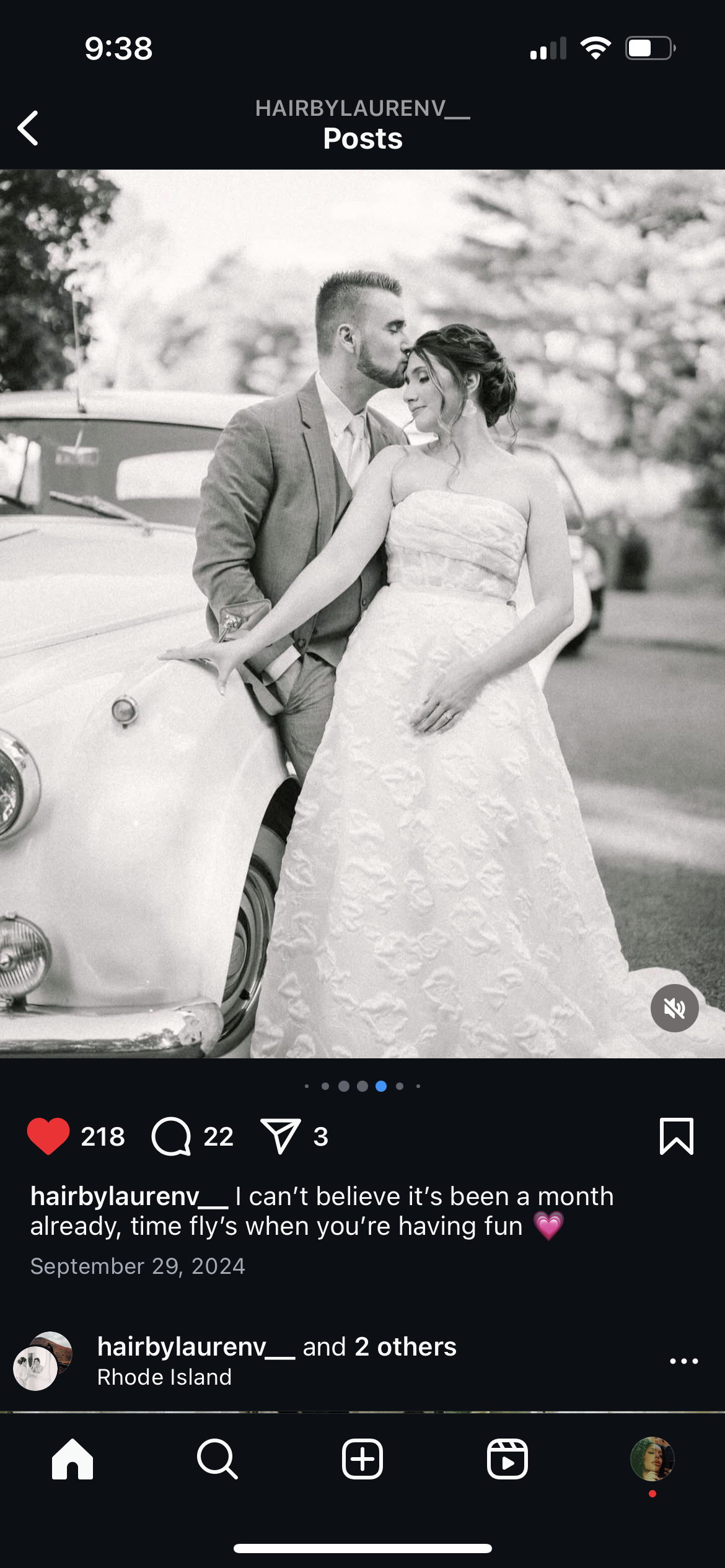 Black and white photo of a bride and groom leaning against a vintage car outdoors, with trees in the background. The groom is kissing the bride on the forehead, and both are smiling gently. The bride is wearing a strapless wedding dress, and the groo