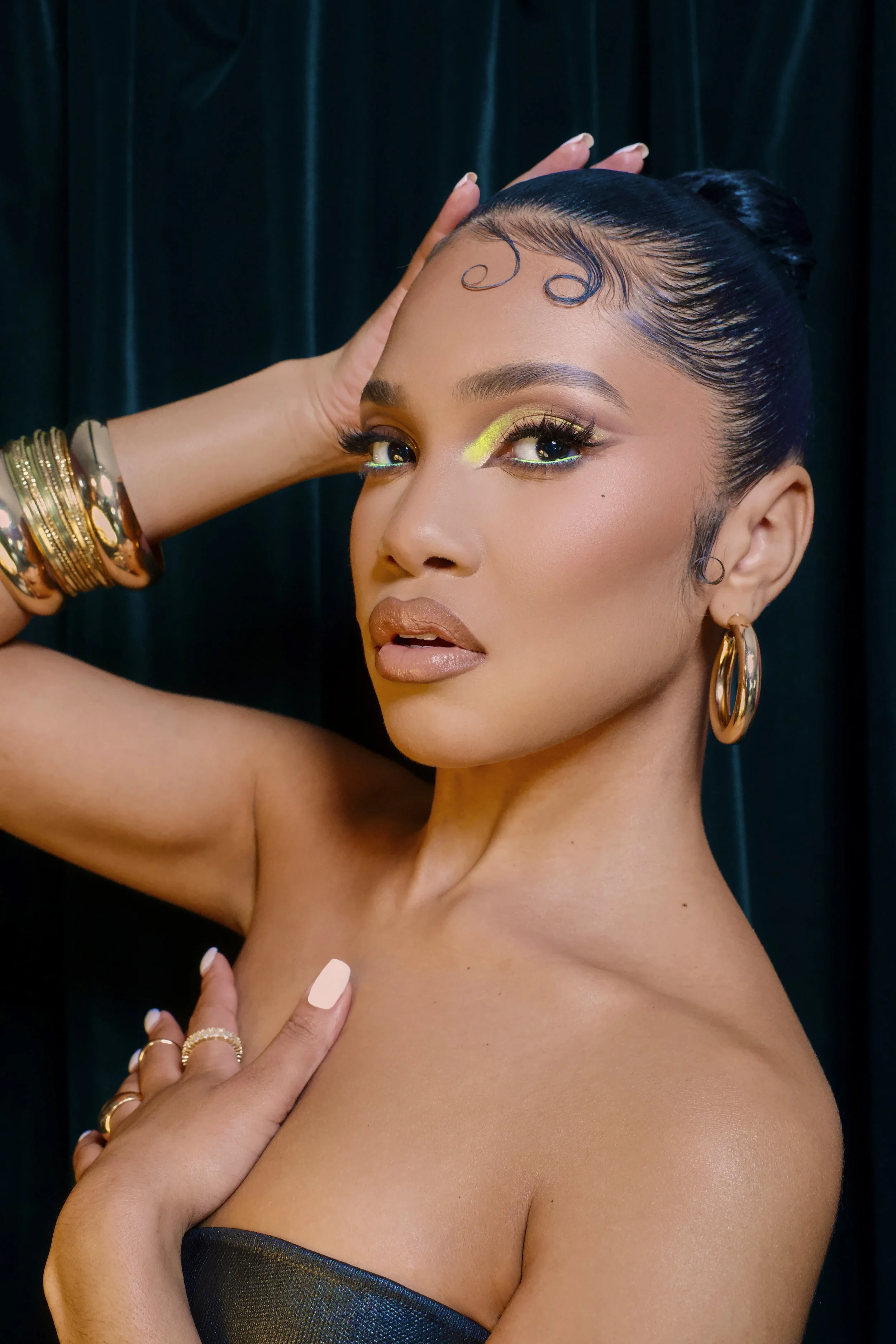 A woman with styled hair, gold earrings, and bracelets, wearing makeup with neon yellow eyeshadow, posing against a dark background.
