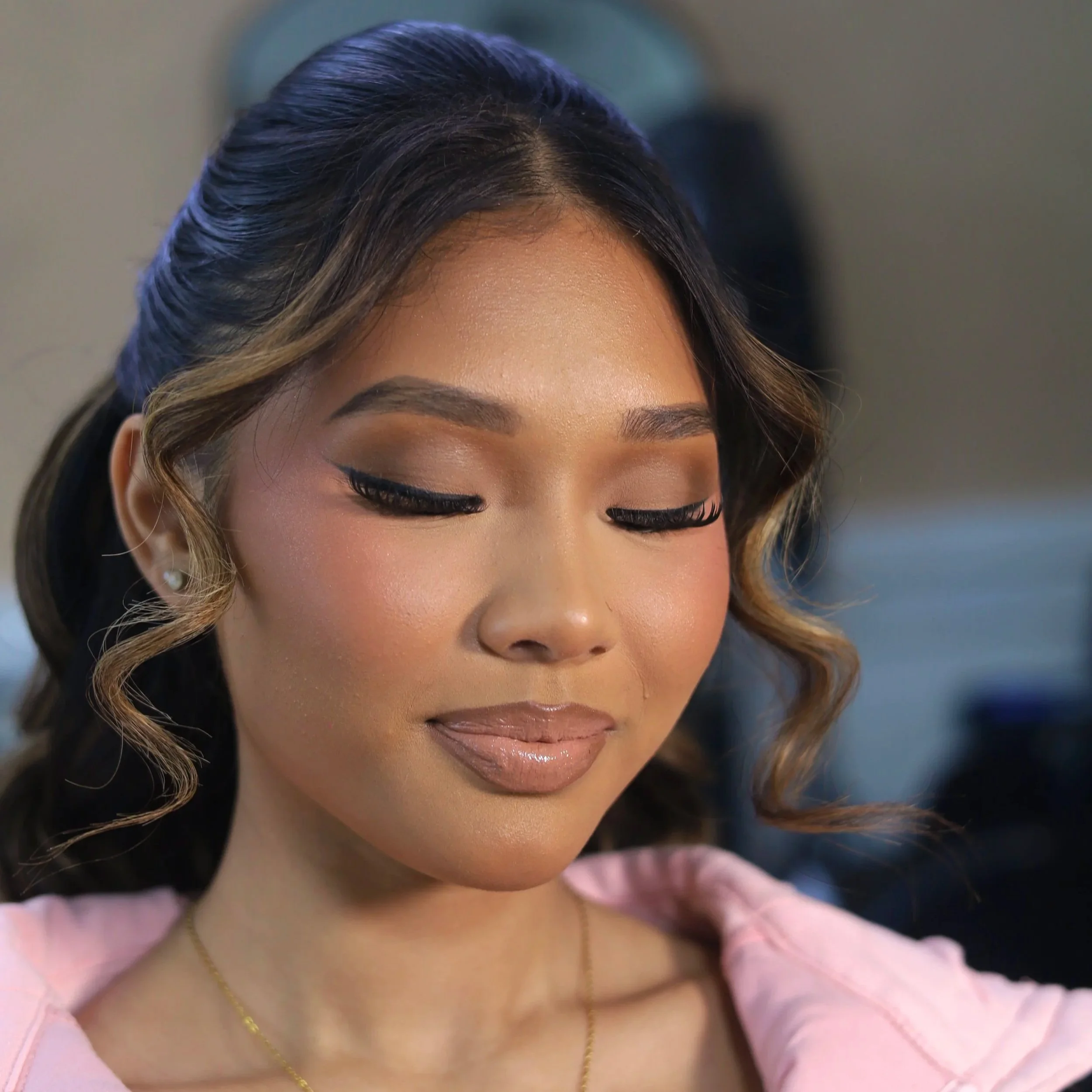 Close-up of a woman with makeup and styled hair, looking downward with eyes closed, wearing a pink top, gold necklace, and small earrings.
