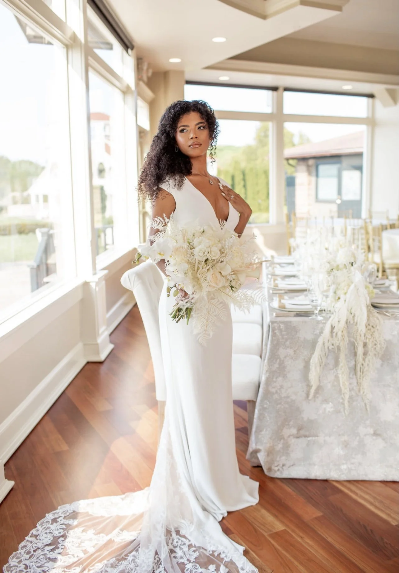 A woman in a white wedding dress holding a bouquet of flowers indoors with large windows and decorated table in the background.
