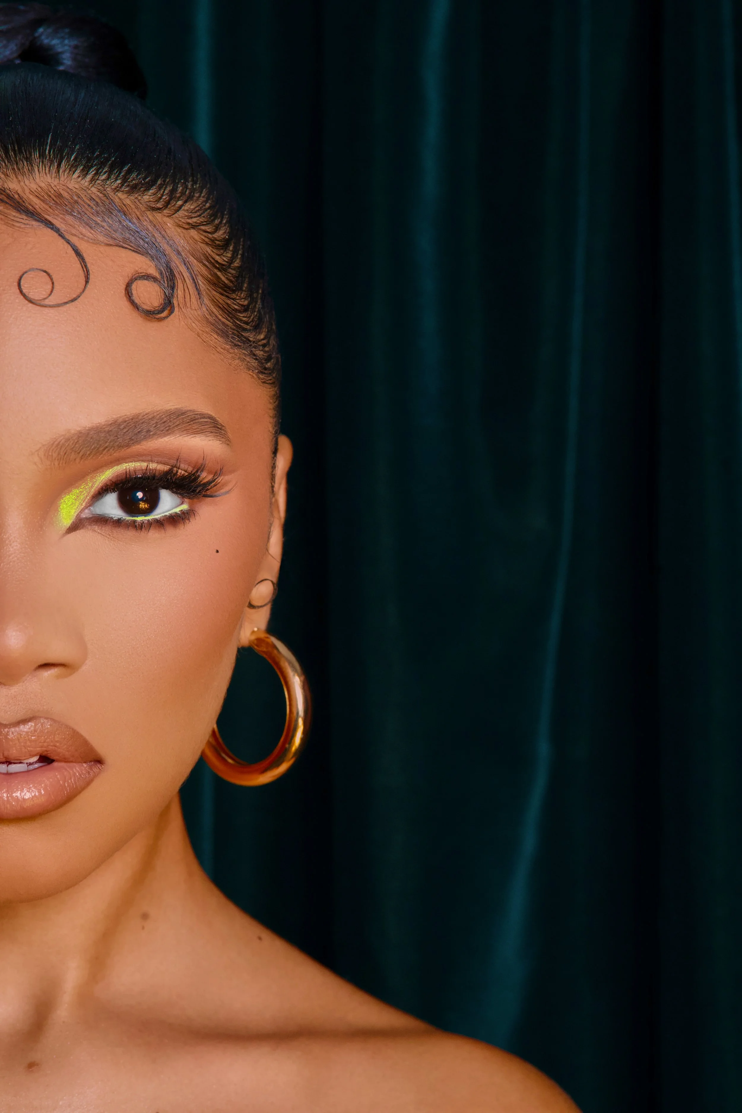 Close-up of a woman with makeup, gold hoop earrings, and styled hair, against a dark teal curtain background.