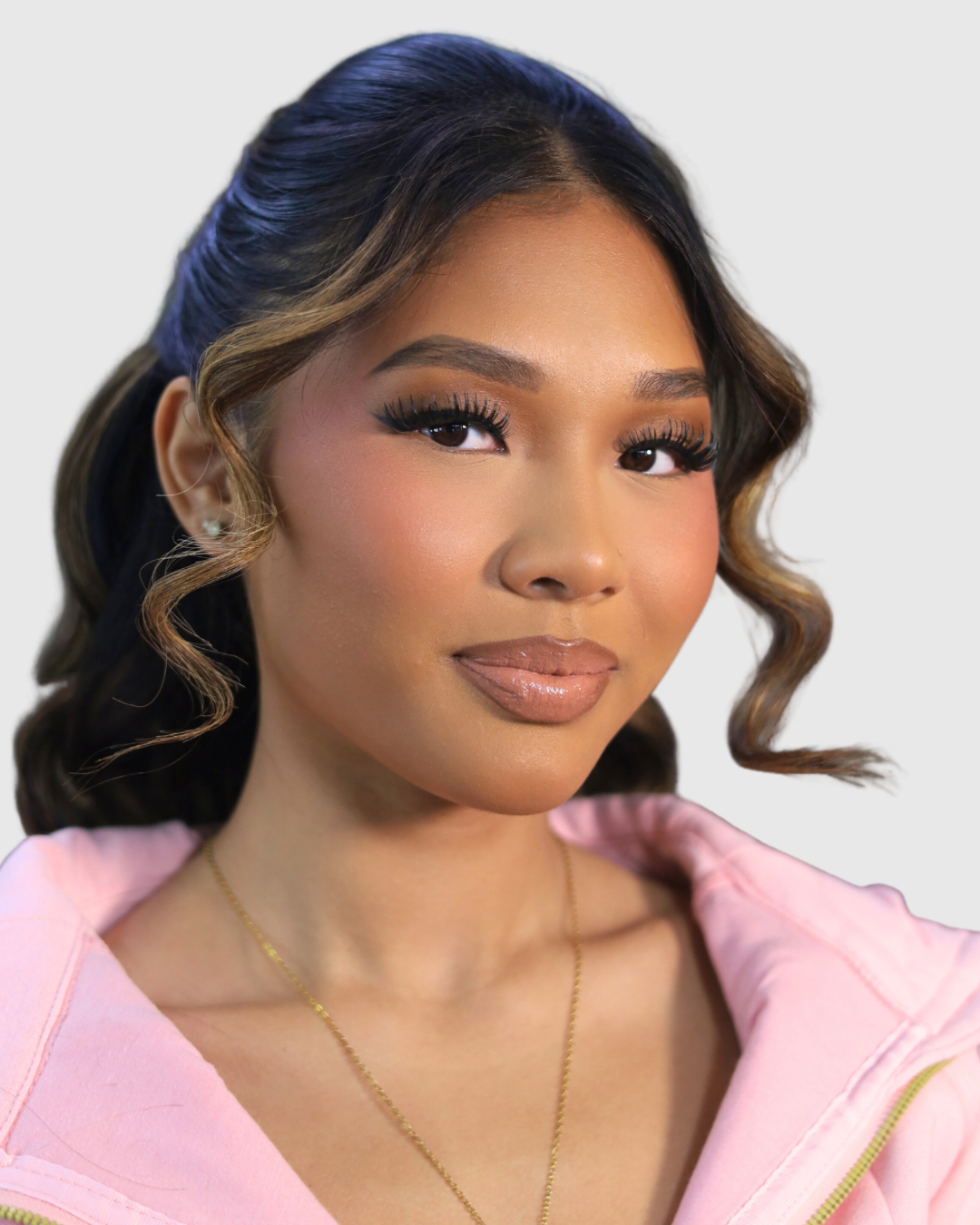 Close-up of a woman with wavy hair wearing makeup, earrings, and a pink top, posing against a plain light background.