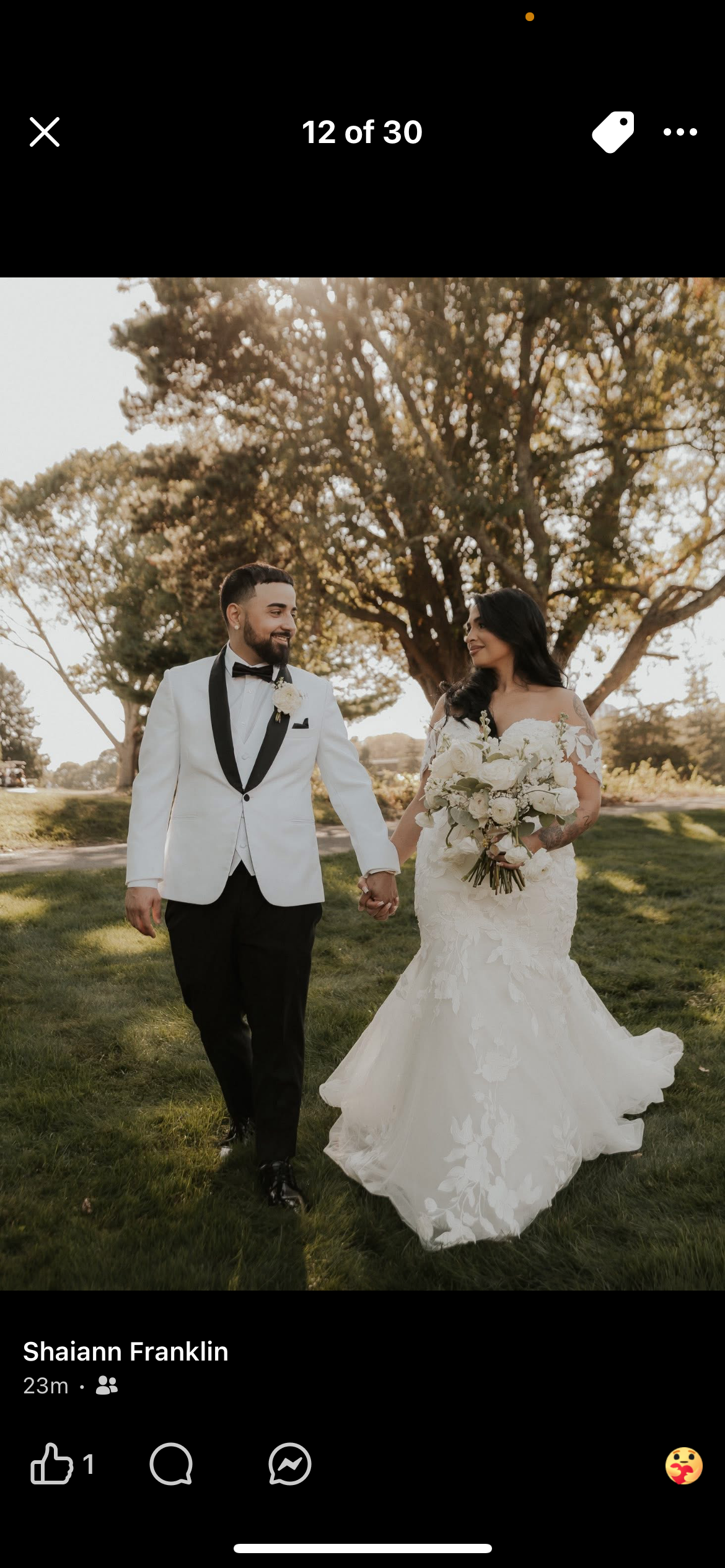 A newlywed couple walking hand in hand outdoors on a grassy area with large trees in the background, the bride holding a bouquet of white flowers, both smiling.