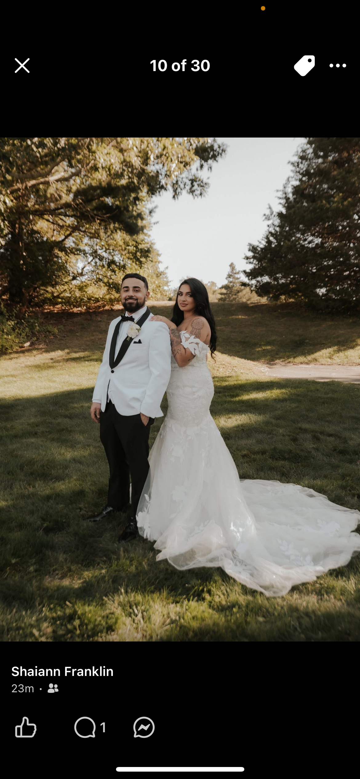 A bride and groom standing outdoors on a grassy area surrounded by trees on a sunny day. The groom is wearing a white tuxedo jacket, black pants, and a black bow tie, while the bride is in a white lace wedding gown with a train. They are smiling and 