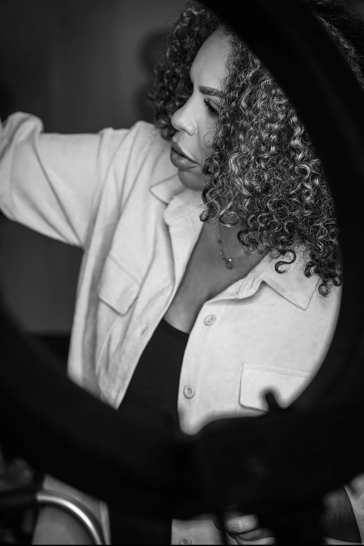 A woman with curly hair wearing a light-colored shirt and a necklace, seen in black and white, looking to the side.