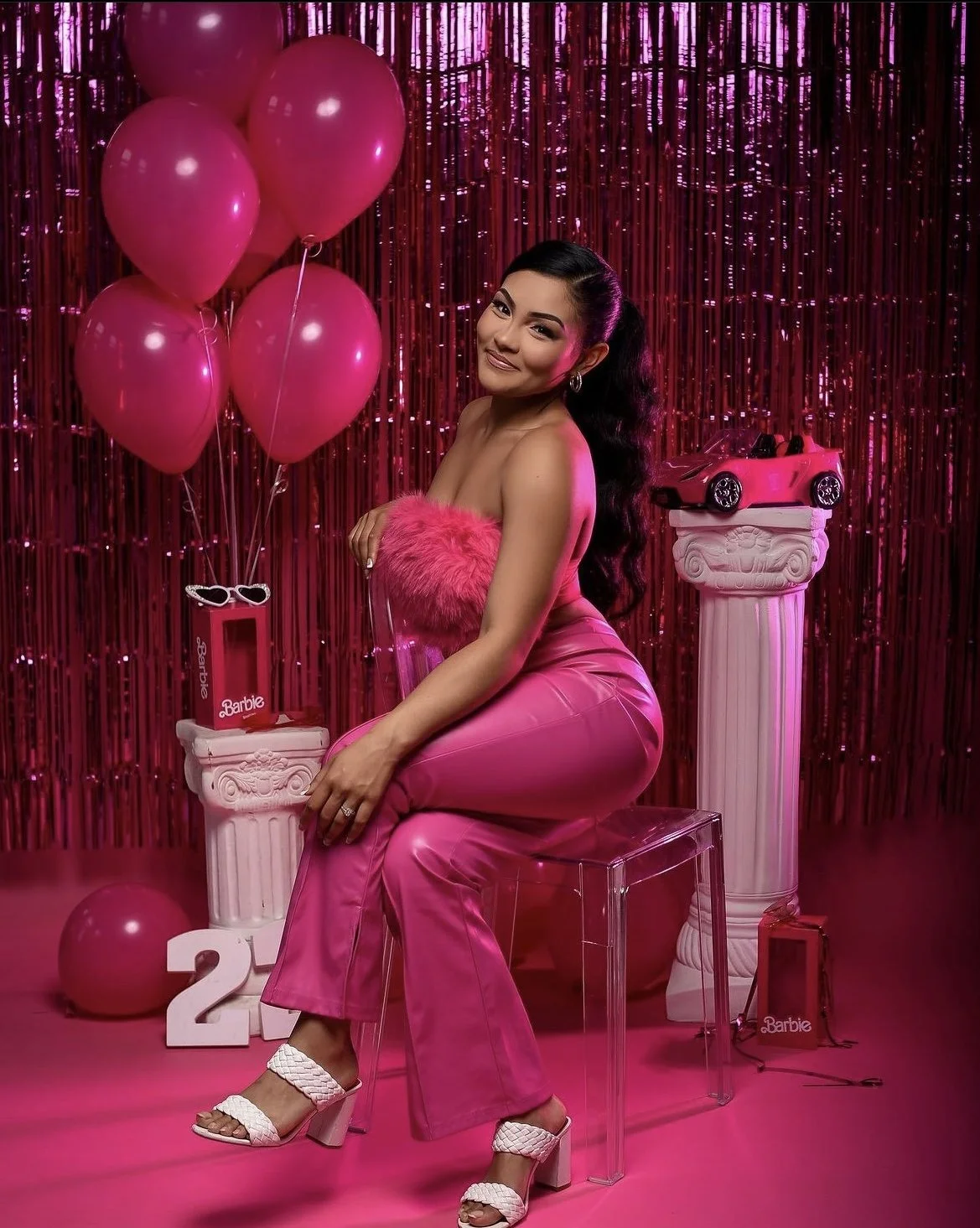 A woman in pink satin pants and a pink fuzzy tube top sitting on a clear chair in front of a pink metallic tinsel backdrop, surrounded by pink balloons, with white decorative columns, a pink Barbie doll, and a pink toy car.