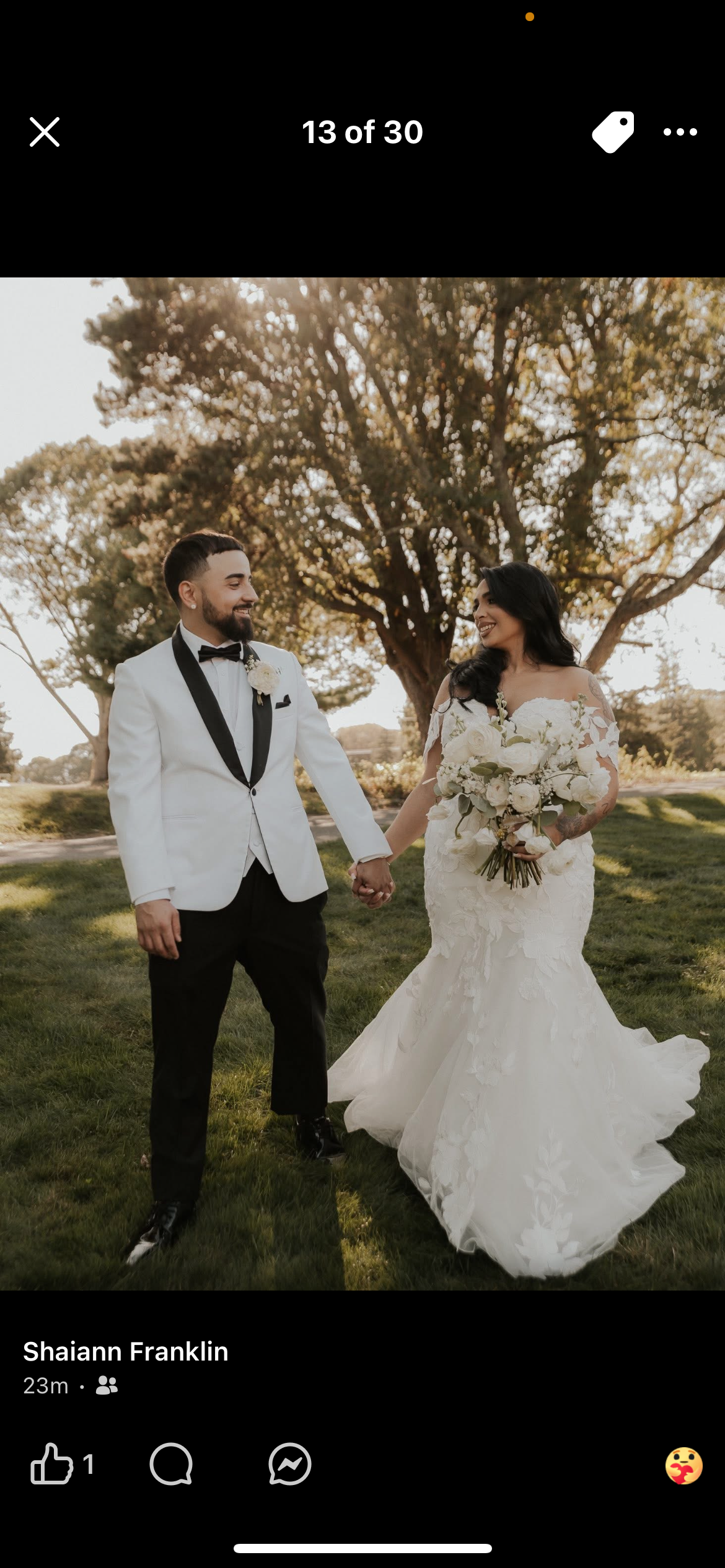 A wedding couple holding hands outdoors under a large tree, with the bride wearing a white lace wedding gown and holding a bouquet of white flowers, and the groom wearing a white tuxedo jacket with black lapels and black pants.
